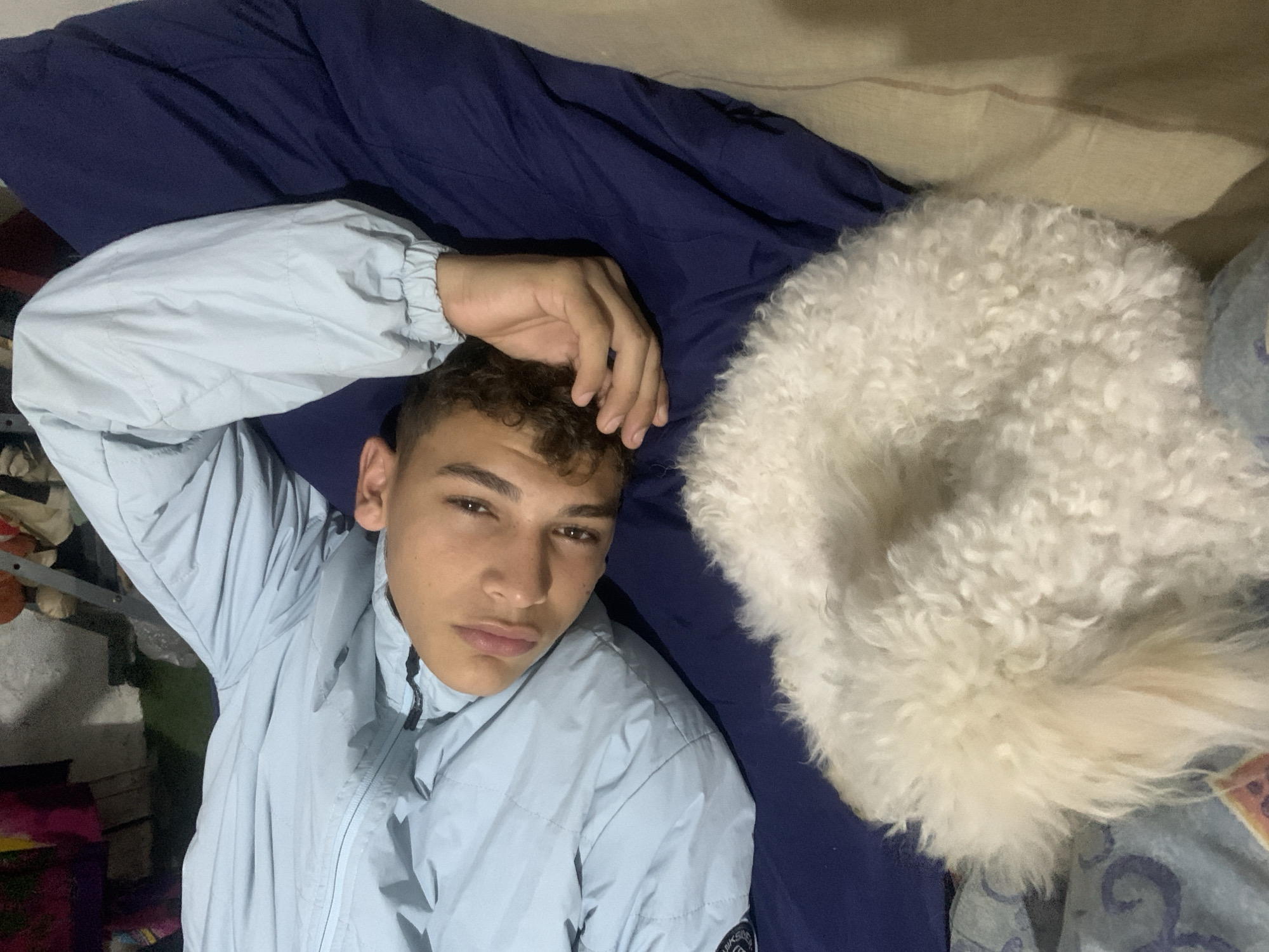 An adolescent male with dark, curly hair lies on his back on a navy blue surface, possibly a bed or couch, wearing a light blue jacket. His right arm is bent with his hand resting near his head. A small, fluffy white dog is curled up on the surface to his left, with its head obscured by its fur. The background is dark and out of focus, but appears to show some items of clothing and possibly a rack or shelf. The lighting is dim and appears to be artificial. The scene is indoors. There is no identifiable information to pinpoint the location in Santa Ana, Venezuela.