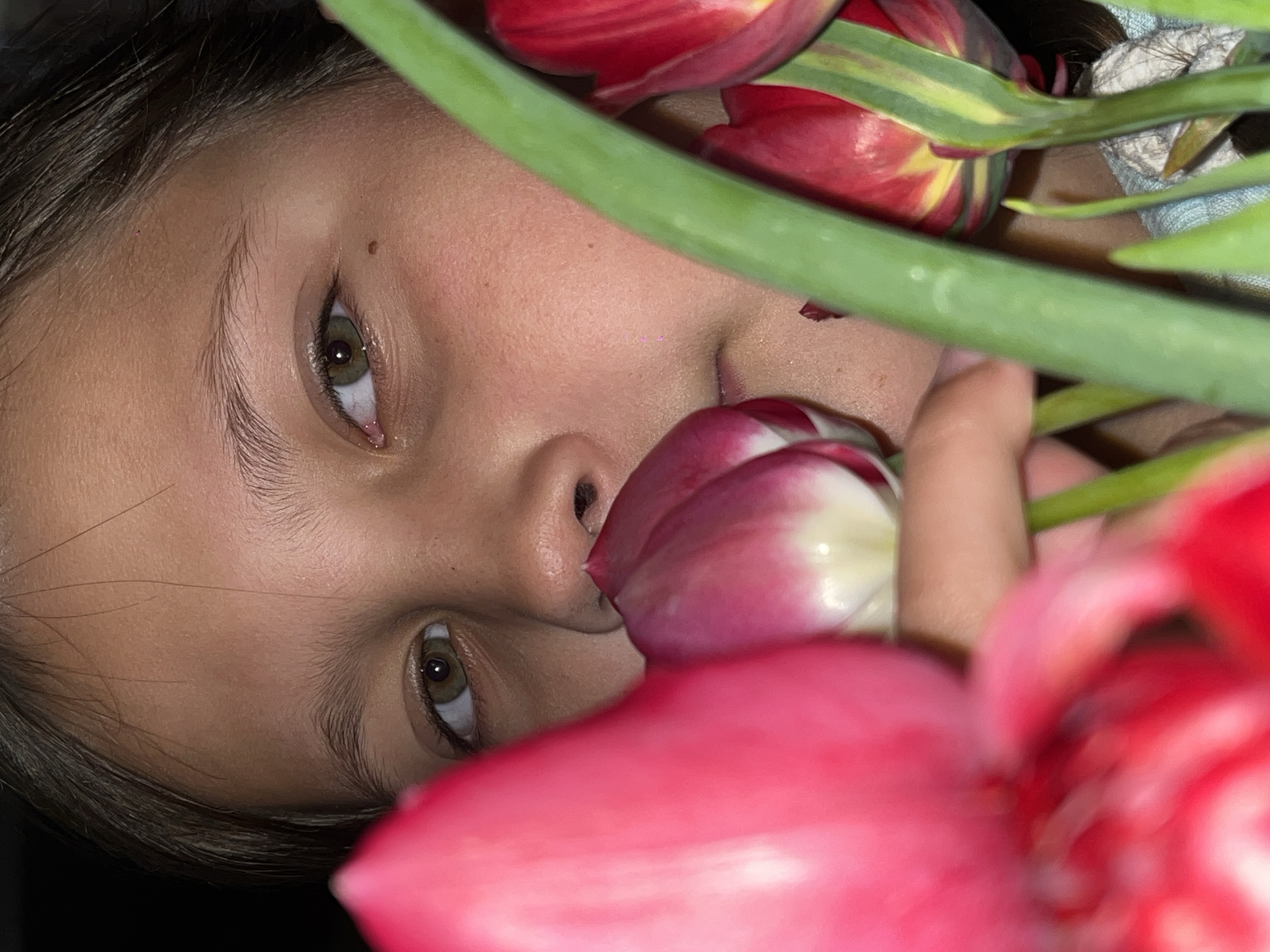 A close-up image features a person's face, possibly a child, surrounded by tulips. The face is angled, with hazel-colored eyes visible. A few freckles are present on the cheek and forehead. The nose appears partially obscured by a red and white tulip. Other red and yellow tulips with green stems surround the face. A hand is partially visible, holding the tulips. The lighting appears natural. There is no indication of the location being Novopokrovskaya, Russia in the image.