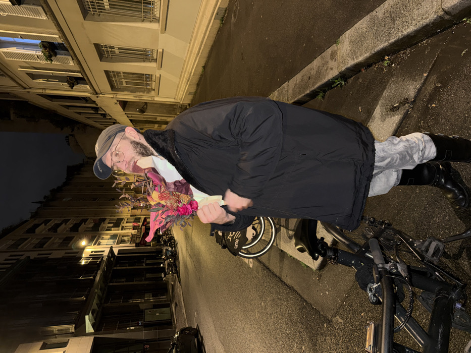A person in a black coat, light gray pants, and black boots stands on a sidewalk, holding a bouquet of flowers. A black bicycle lies on its side beside them. The setting appears to be an urban street at night, with buildings visible in the background. The sidewalk is adjacent to a road. The lighting suggests it is evening.