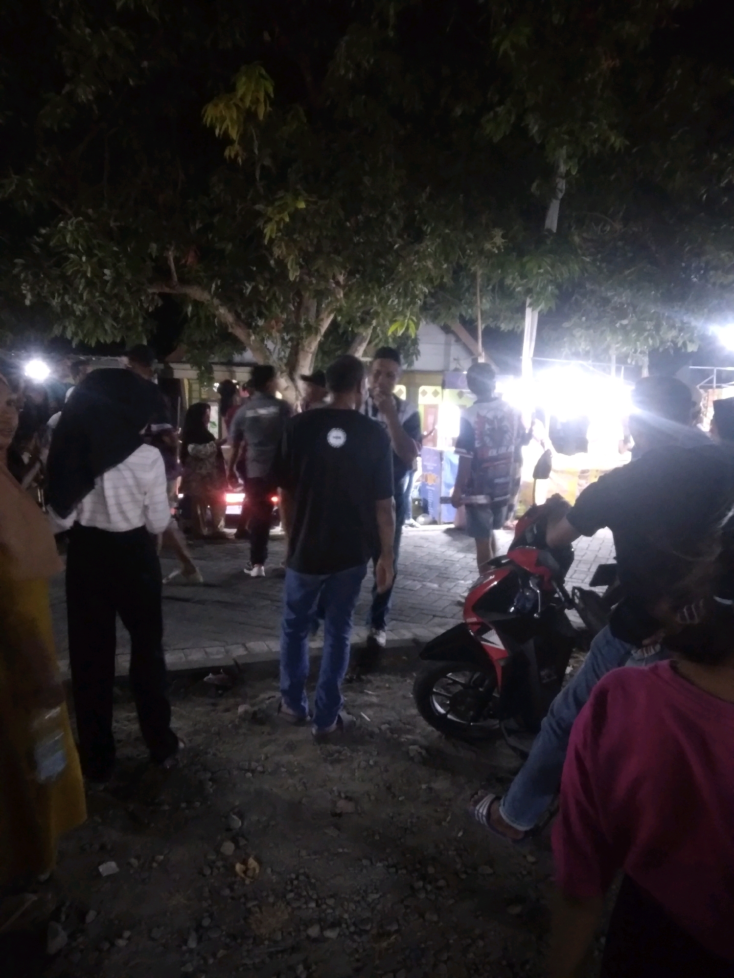A nighttime scene in Watestanjung, Indonesia, depicts a group of people gathered outdoors, possibly at a market or event. Several individuals are standing and walking around. In the foreground, a red and black motorcycle is parked. The ground is covered in gravel and debris. Trees with lush foliage are visible in the background, along with some illuminated stalls or tents. The lighting suggests artificial illumination from nearby sources.