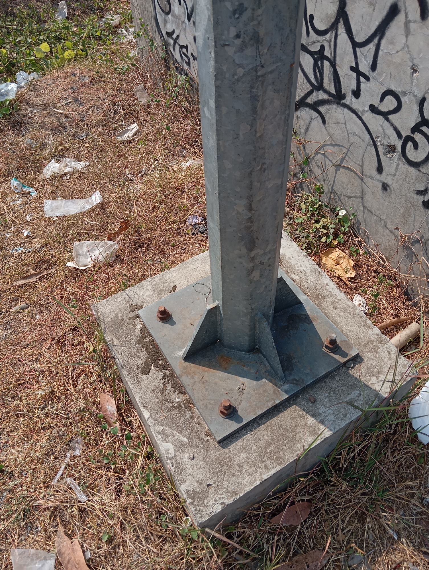 The image shows the base of a metal pole, likely a lamppost or utility pole, anchored to a concrete foundation. The pole and its base plate are made of galvanized metal, with visible bolts securing it to the concrete. The area surrounding the pole is covered in dry, brown grass and scattered debris, including plastic wrappers and leaves. A concrete wall, marked with black graffiti, forms the background. The scene suggests an outdoor urban environment, possibly a roadside or public space. The image was taken in Sokaraja, Indonesia. There are no people or animals visible in the image.
