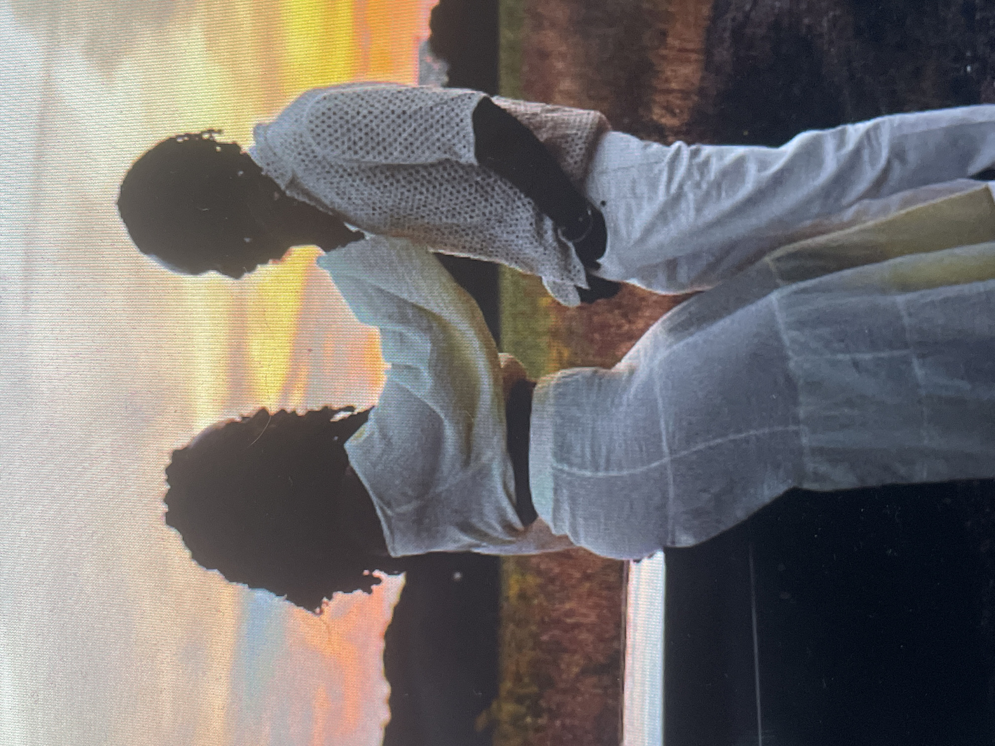 The image, when viewed in its correct orientation, captures two individuals in silhouette against a vibrant sunset or sunrise sky. The scene is outdoors, bathed in strong backlighting that creates dark, undefined profiles of the subjects while illuminating the sky with rich, warm colors.
Two adults are depicted, standing closely and facing each other in what appears to be an intimate or affectionate moment. The person on the left wears a light-colored, possibly white, collared shirt with a distinct textured or knit pattern, and matching light-colored pants. They are leaning in, with their head slightly bowed and their left arm raised, possibly touching or holding the other person. The individual on the right is adorned in a flowing, light-colored dress or a coordinated skirt and top. Their arm is extended towards the other person as they gaze upon them. Both figures have dark, possibly curly, hair; one appears to have shorter hair and the other longer.
The background is dominated by a dramatic sky, painted with gradients of orange, yellow, and soft red, characteristic of either dawn or dusk. The landscape beyond the figures is dark and indistinct, suggesting distant hills or dense foliage. A dark, rectangular element is visible at the bottom right, possibly part of a structure like a railing or bench. The pose and proximity of the individuals convey a sense of tenderness, connection, or romance.
The image quality suggests it may be a photograph of a digital screen, evidenced by the visible horizontal lines and slight pixelation. No text is visible within the image. While the context specifies Sofia, Bulgaria, there are no distinct visual cues within the photograph itself to confirm this location.