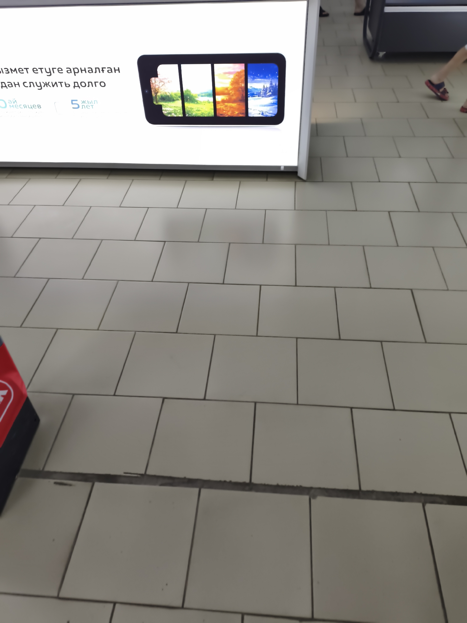 The image shows a display advertising a product with text in what appears to be Kazakh. The text includes "ай месяцев" (months) and "5 жыл" (5 years). The display features a tablet-like screen divided into four sections, each showing a different season: spring, summer, autumn, and winter. The background of the display is white. To the right of the display, a person is walking away, wearing red sandals. The floor is tiled with square, light-colored tiles. In the lower left corner, a red and black object, possibly a shopping cart or a display stand, is partially visible. The scene suggests a commercial or public indoor space, possibly a mall or store, in Aktobe, Kazakhstan.