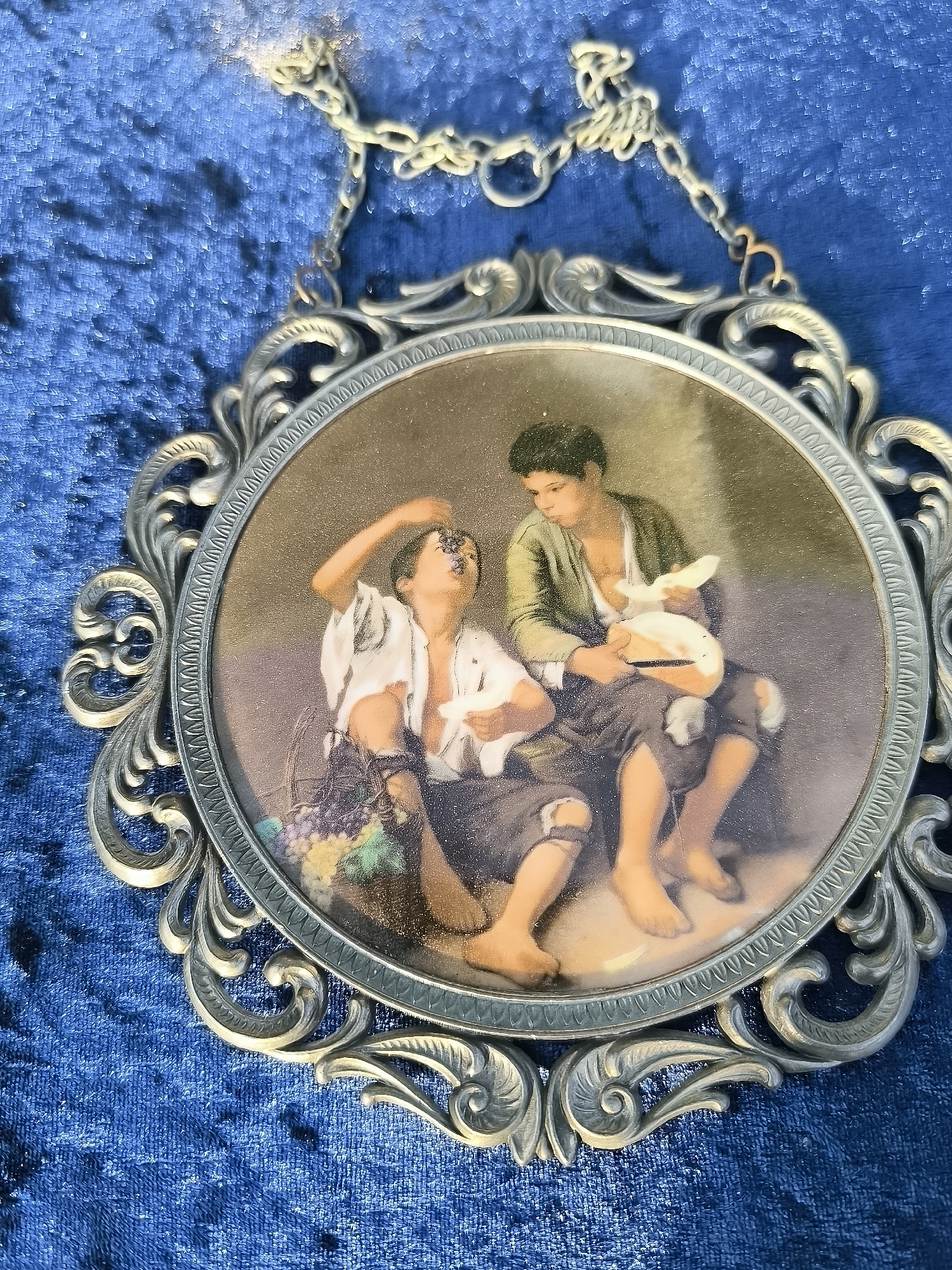 The image shows a circular framed artwork depicting two young boys. The boy on the left wears a white shirt and dark pants, with a basket of grapes at his feet. He is shown reaching up to eat grapes. The boy on the right wears a green shirt and dark pants, holding a loaf of bread. Both boys are barefoot. The artwork is hung from a chain on a textured blue surface, possibly velvet. The frame is ornate and metallic. The location of this item within the city of Klaipėda, Lithuania, is not ascertainable from the provided image.