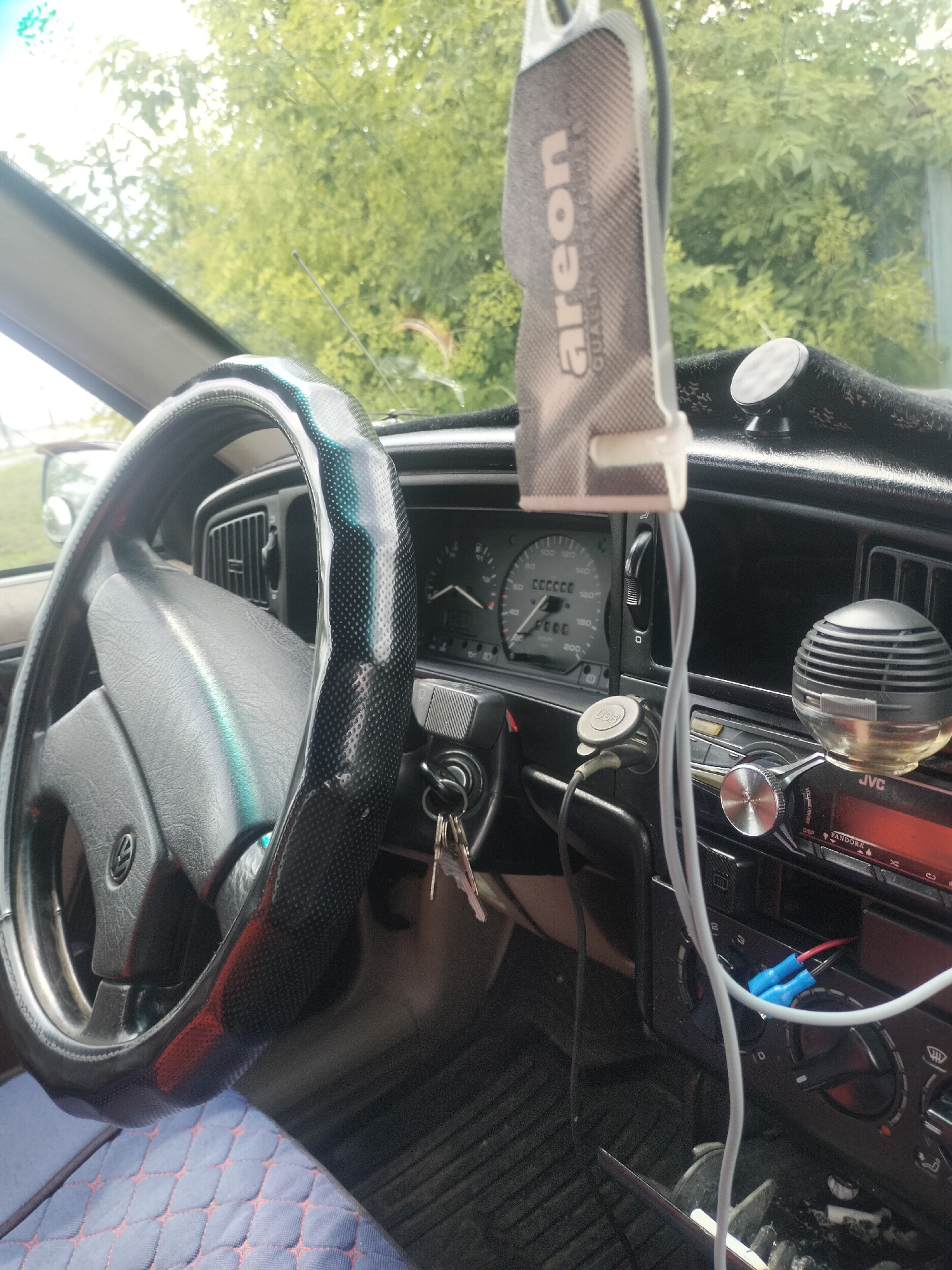 The image shows the interior of a car, likely parked outdoors. The dashboard is visible, featuring a steering wheel with a textured cover, a speedometer, and a car stereo. An air freshener hangs from the rearview mirror. The view through the windshield shows green foliage, suggesting a natural or suburban setting. The image does not provide any information that would allow for its location in Кіші Чураковка, Kazakhstan.