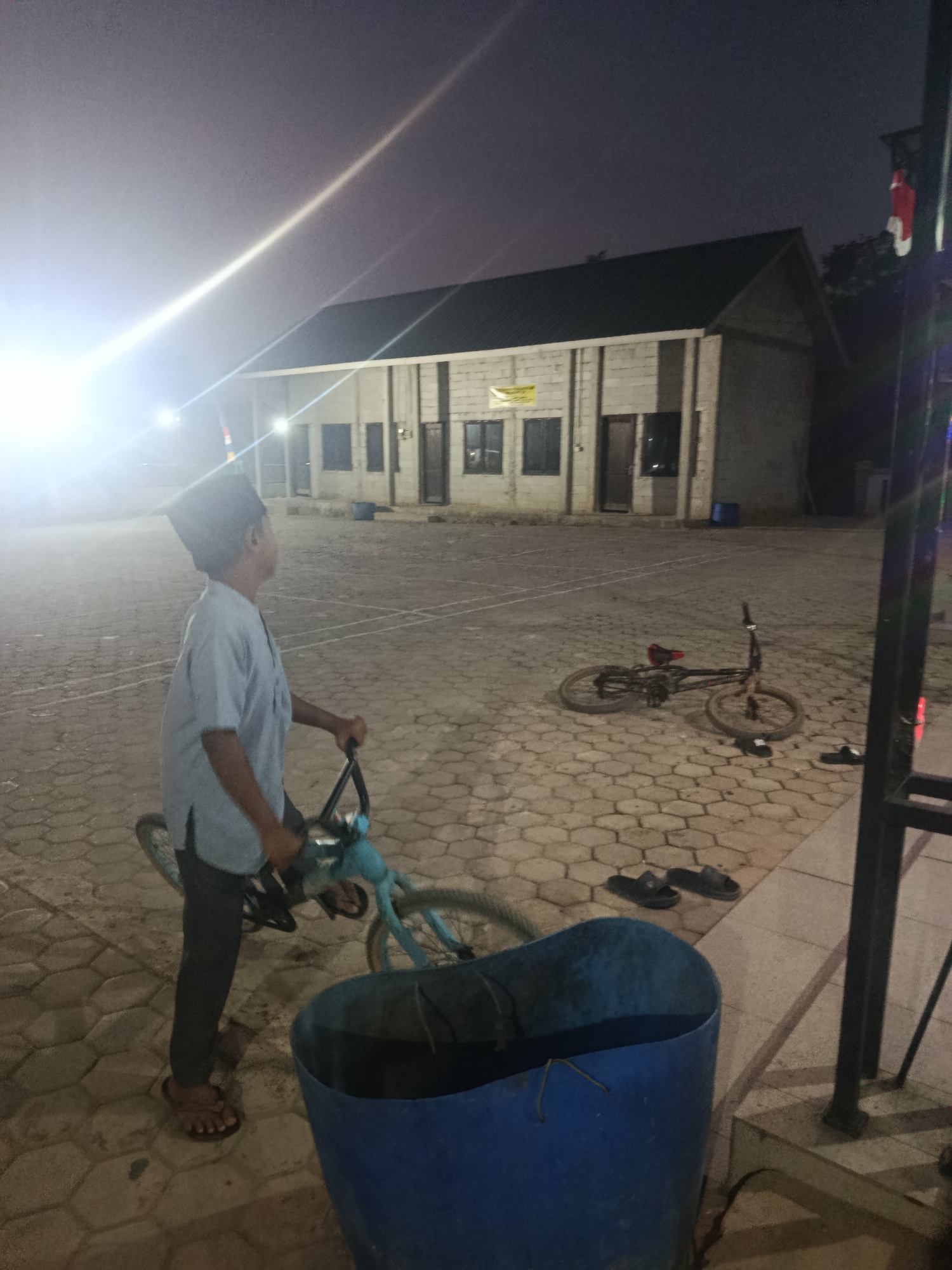 In Swarga Bara, Indonesia, an outdoor scene unfolds at dusk. A young boy, wearing a blue shirt and dark pants, is positioned to the left of the frame, looking to his right while holding onto the handlebars of a light blue bicycle. The bicycle is partially visible. To the right of the boy, a black bicycle is lying on its side on a paved ground made of interlocking hexagonal stones. In the background, a light-colored building with a dark roof stands. The sky above is a muted purple. In the foreground, a large blue trash can is present. Further to the right, a black metal railing is visible. The overall lighting suggests it is evening or night, with artificial light sources creating bright flares.