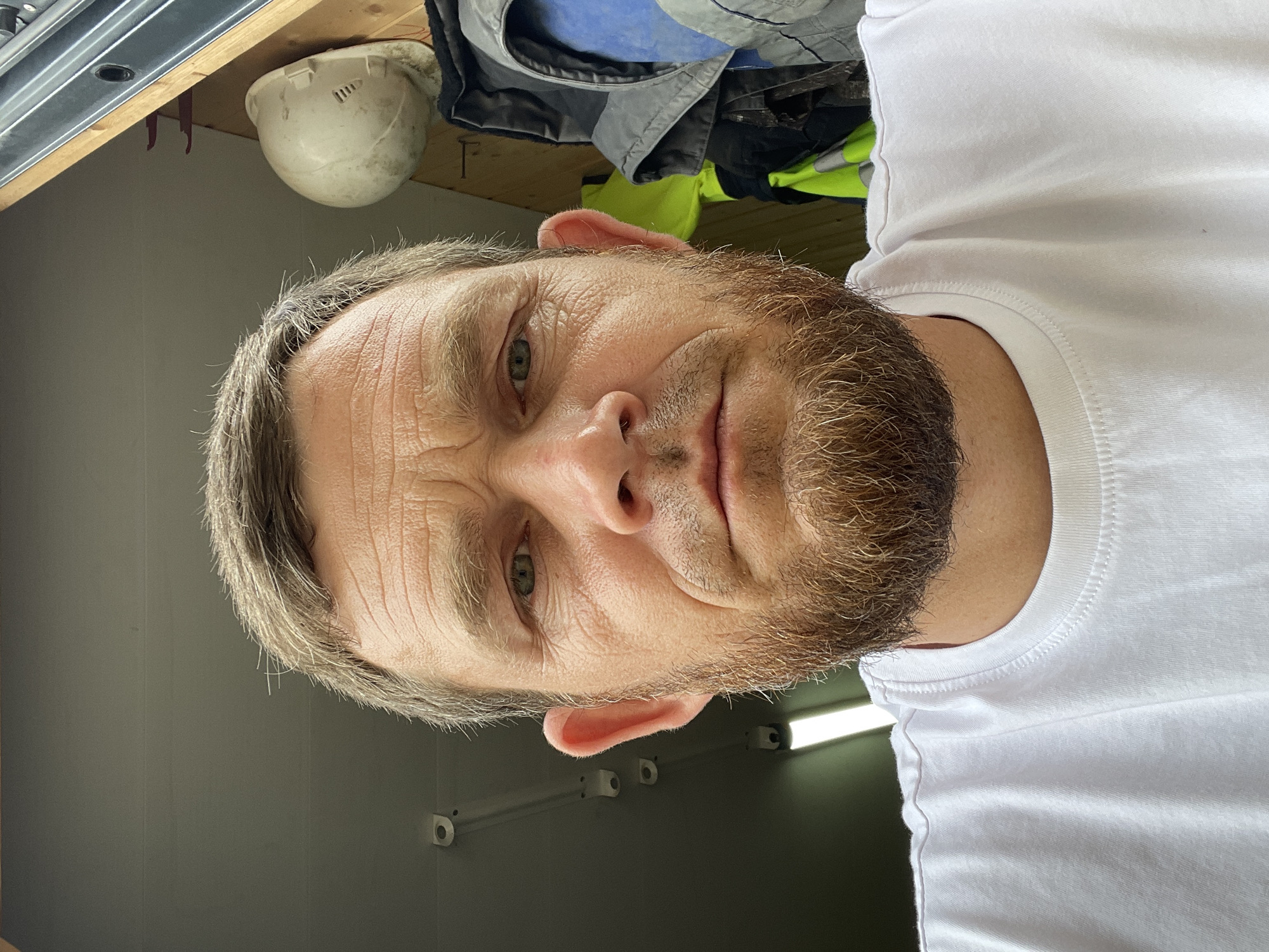 The media file is a close-up, selfie-style portrait of a man in a white t-shirt. The man has light skin, graying hair, a beard, and green eyes. He appears to be looking directly at the camera with a neutral expression. His face is wrinkled, suggesting middle age.
In the background, to the left of the man's head, there is a white hard hat resting on a wooden surface, possibly a shelf. Behind the hard hat, there are pieces of clothing, including what appears to be a high-visibility jacket in yellow and gray, and a blue garment. The setting looks like an indoor space, perhaps a workshop, garage, or storage area, given the presence of the hard hat and workwear. A metal track is visible at the top left of the image, and a fluorescent light fixture is visible in the background to the right. The wall appears to be a neutral gray color.
There is no visible text in the media file. The lighting suggests it is daytime, though the interior setting makes it difficult to ascertain specific time of day or weather conditions. The overall context suggests a personal moment captured, possibly during or after work, given the work-related items in the background. The image is oriented sideways, with the man's head at the top of the frame.