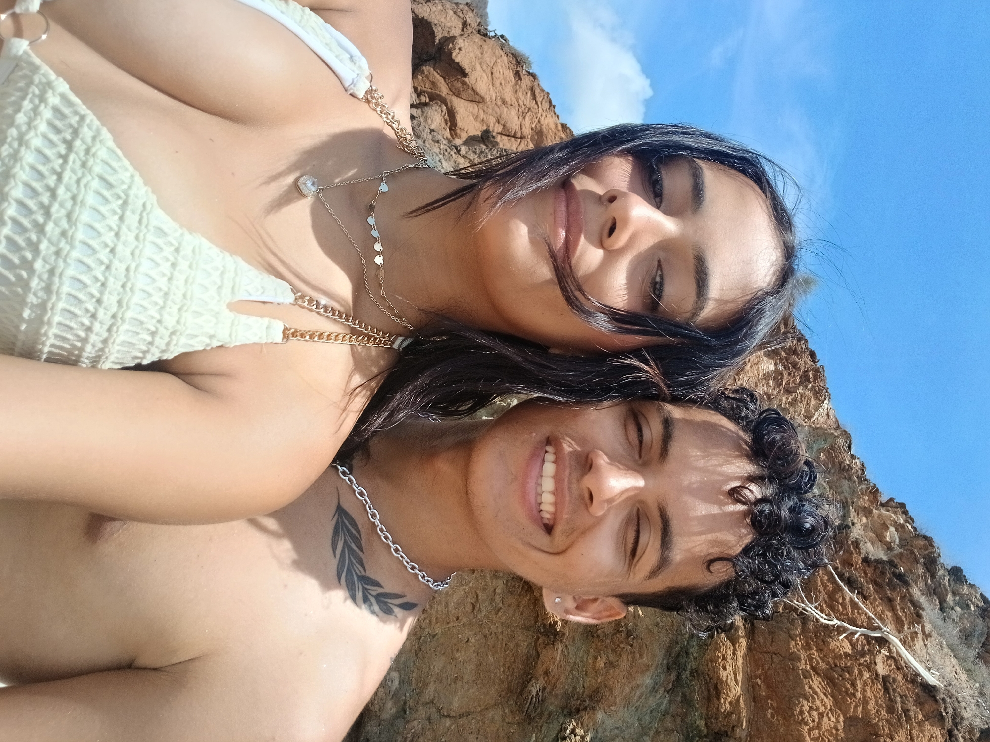 This close-up image, taken in Juan Griego, Venezuela, features two individuals, a woman and a man, in a rotated selfie-style shot, suggesting they are lying down or leaning back. The perspective is looking upwards towards the subjects and the sky.
The woman is positioned in the upper part of the frame, smiling subtly. She has dark hair and wears a white, textured bikini top with golden chain details. Layered gold necklaces adorn her neck; one features a sparkling, presumably jeweled, pendant, and another has small, flat circular charms.
The man is visible below and slightly behind the woman, sporting a broad, bright smile that reveals his teeth. He has dark, curly hair and a tattoo of a stylized leaf or feather design on his left shoulder/neck area. A simple silver chain necklace and a small ear piercing are also visible.
The background reveals a rugged, brownish-orange cliff face, indicating an outdoor, possibly coastal or mountainous, setting. Above the cliff, a clear, vibrant blue sky with a few wispy white clouds confirms bright, sunny weather. Strong sunlight casts shadows and highlights on the subjects, emphasizing their relaxed and happy expressions. There is no visible text in the image.