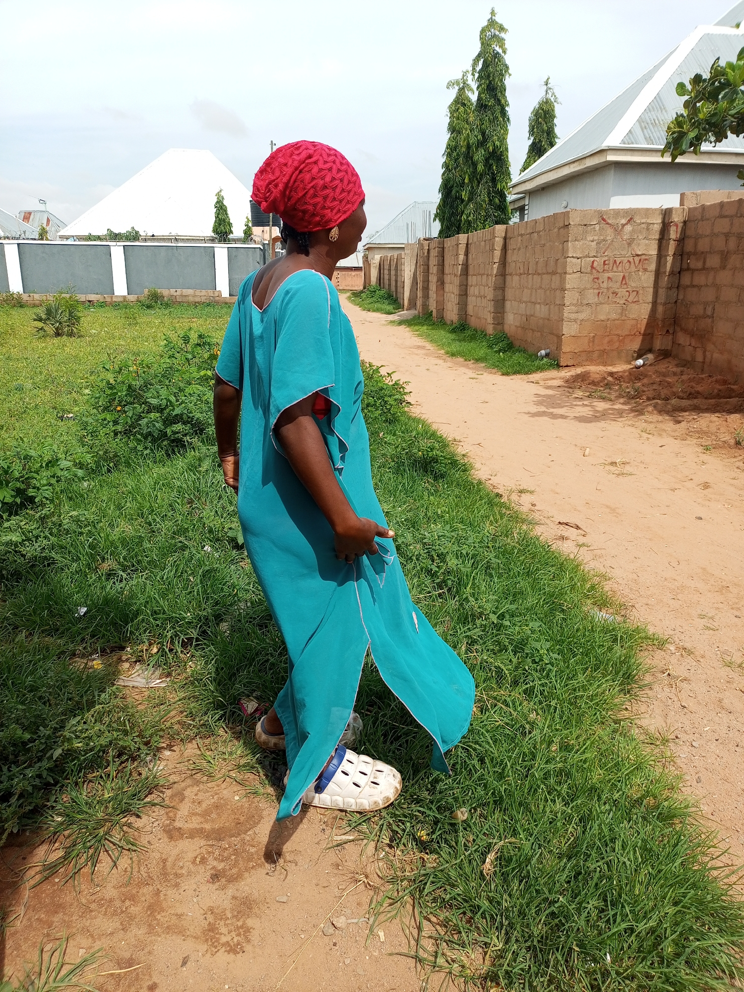 In Tempure, Nigeria, a woman stands on a dirt path beside a patch of green grass, looking away from the camera. She is wearing a turquoise abaya and a red headscarf. The abaya has white trim on the sleeves and a slit in the side. She is also wearing white slip-on shoes. Behind her, a brick wall runs alongside the path. Beyond the wall are houses with metal roofs and some tall trees. The sky is cloudy. There is a white conical building in the background.