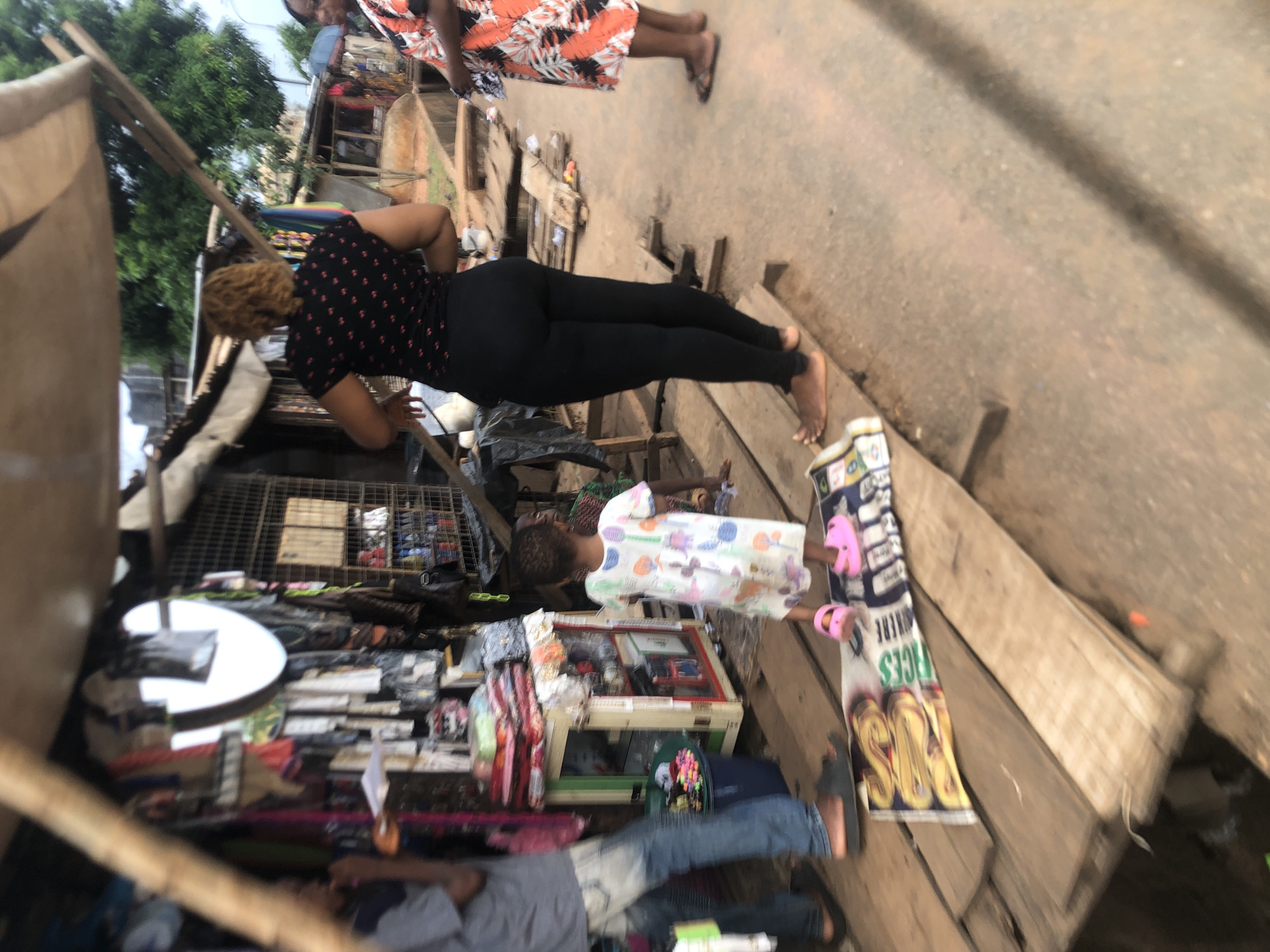 The scene is located in Ukwu-Abwa, Nigeria, appearing to be a street market environment. A woman in a black shirt with red flowers and black leggings is interacting with something behind a display. A child in a white dress with printed patterns is standing. A person in a floral dress is walking down the street. There is a sign advertising shoes/slippers. The background contains a cluttered market stall with a variety of goods and people. The street is paved, but in poor condition.