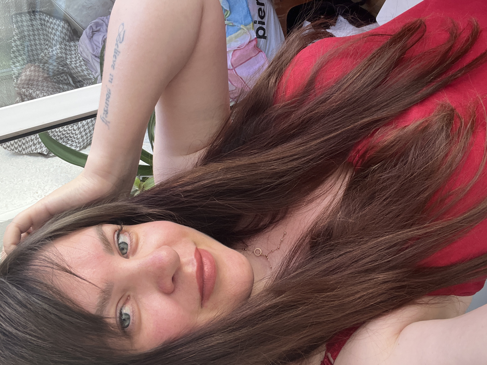 The media file shows a close-up, overhead shot of a young woman lying down. She has long, dark brown hair that is spread out around her. Her eyes are open and blue-green, looking towards the camera. She is wearing a red top and a delicate necklace with a small circle pendant. On her left forearm, a tattoo with the words "Believe in yourself" is visible.
The background is blurred but suggests an indoor or semi-outdoor setting. Hints of patterned fabric and what appears to be a window with a view of greenery are discernible. The lighting suggests it is daytime. The woman's expression is relaxed and direct. There is no specific event or activity captured, only a posed portrait. The setting is identified as Noviy, Russia.