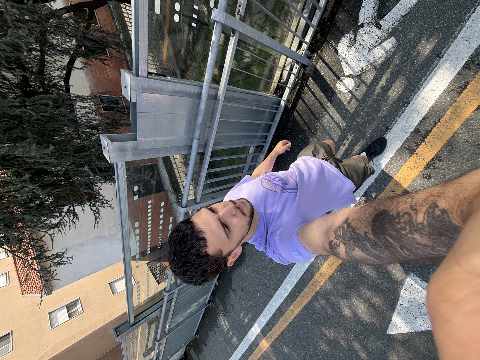 An overhead-angle selfie captures a young man standing on what appears to be a pedestrian bridge or overpass in Vigevano, Italy. The man, with dark hair and a beard, looks towards the camera with a neutral expression. He wears a light purple t-shirt with "LOS ANGELES" text and graphics visible, dark shorts, and black sneakers. A large, dark, wave-like or abstract tattoo covers a significant portion of his right leg, and another smaller tattoo is visible on his left forearm.
The setting is outdoors during bright daylight, casting distinct shadows from the metal railing of the overpass onto the asphalt below. The ground features white and yellow road markings, including a partial white arrow and a solid white line. To the left, lush green trees partially obscure buildings, one of which is reddish-brown with white window frames and another lighter beige/brown structure with multiple rectangular windows and tiled roofs, characteristic of European architecture. The metal railing of the overpass, constructed from galvanized steel, extends across the scene, providing a barrier. The overall impression is an urban or suburban environment with a clear, sunny day.