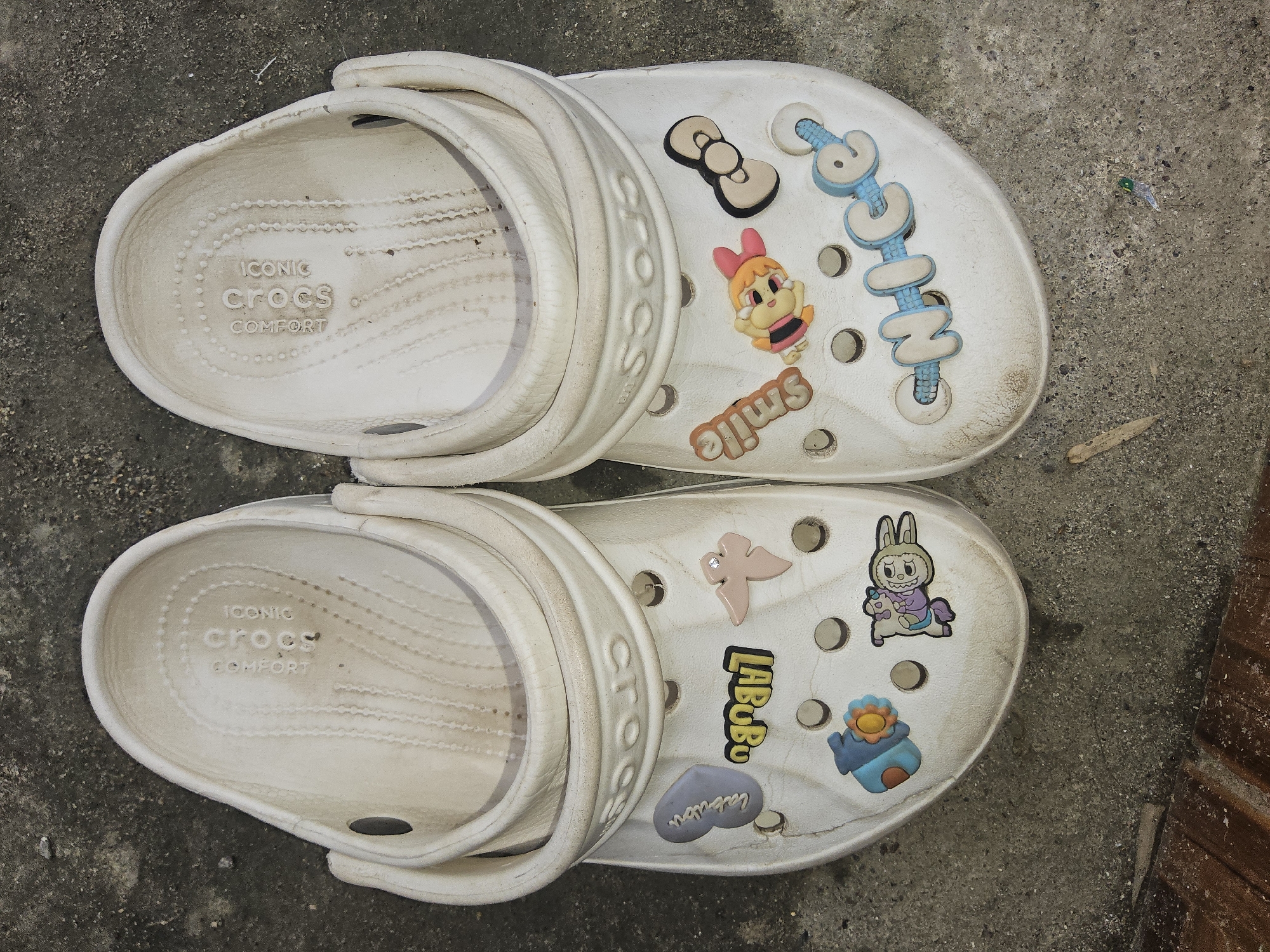 The image shows a pair of white Crocs shoes with various colorful Jibbitz charms attached to them. The shoes are placed on a textured concrete surface, possibly a sidewalk or ground in an outdoor setting. The Jibbitz include a blue heart with cursive writing, the word "LABUBU" in yellow and black, a small pink bird with a diamond accent, a blue house with an orange flower, a rabbit character, the word "Smile" in orange, a cartoon character with pink hair and a bow, a black and tan bow, and the word "CHICE" in blue and white letters. The shoes appear worn and slightly dirty. The concrete surface is speckled with various shades of gray and appears to be a public area, consistent with an urban or semi-urban environment in Payagaboh, Indonesia. There are no people or animals visible in the image, and no actions or interactions are depicted.
