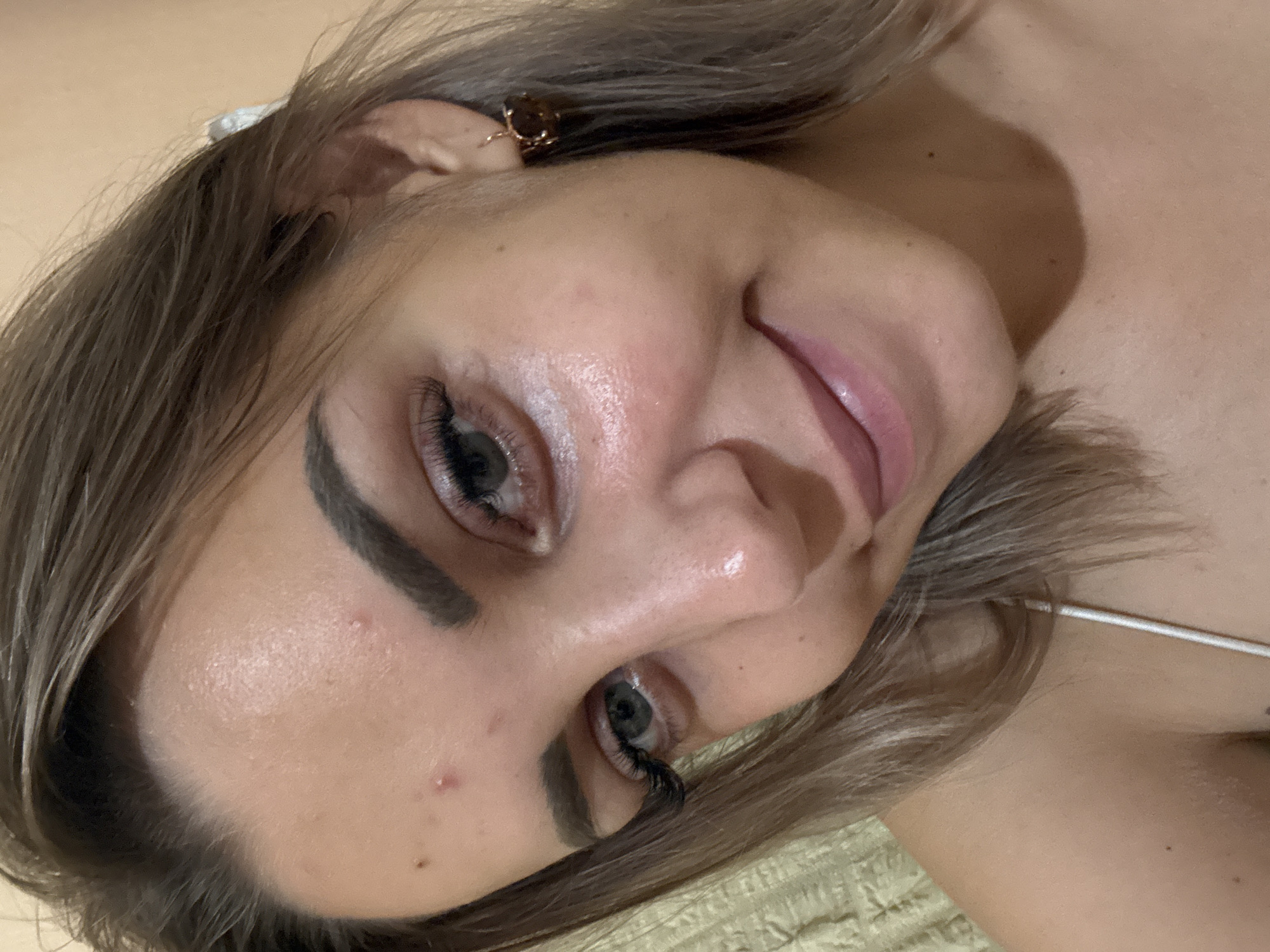The image shows a close-up, head-and-shoulders shot of a young woman with light brown hair and green eyes. She is wearing makeup, including mascara and lip gloss, and has on a small, rose-gold colored earring with a dark stone. There are a few blemishes on her forehead. She is looking directly at the camera with a slight smile. The background is out of focus and appears to be a wall with a peachy hue. The image does not contain any information that would allow for the determination of its location in Kholmsk, Russia.