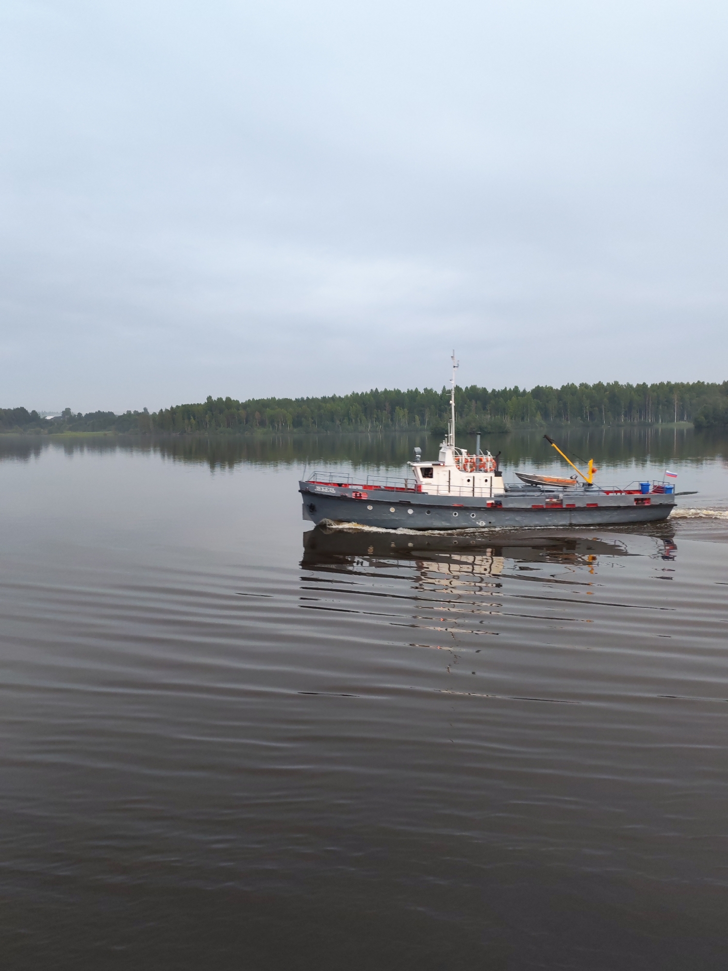 A medium shot captures a gray and red tugboat moving across a calm, dark body of water under an overcast sky. The boat is angled to the right and is leaving a white wake. It has a white superstructure with windows and a red railing around the deck. A small wooden boat is secured on its deck, as is a yellow crane. A Russian flag is visible at the stern of the tugboat. In the background, a line of dense green trees borders the water, and a hazy, distant shoreline is visible to the left. The water's surface is rippled, creating dark and light patterns. The scene appears to be in a natural setting, possibly a river or lake, rather than a city. No people are visible on the boat or in the surrounding environment. The image does not provide any specific information about the location in Ratibor, Russia, as Ratibor is a city in Poland, not Russia, and the image depicts a natural landscape.