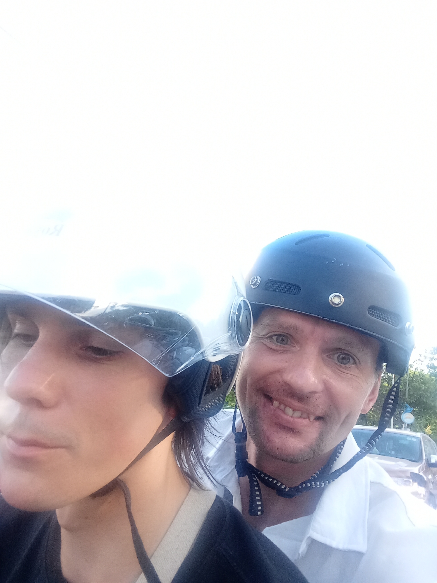Two people wearing helmets are on a scooter in Da Nang, Vietnam. The person in the foreground is on the left, facing left, with a clear visor partially obscuring their face. The person behind them, on the right, is smiling at the camera, wearing a dark helmet and a white shirt. In the background, a car and some trees are visible. The lighting is bright, suggesting daytime.