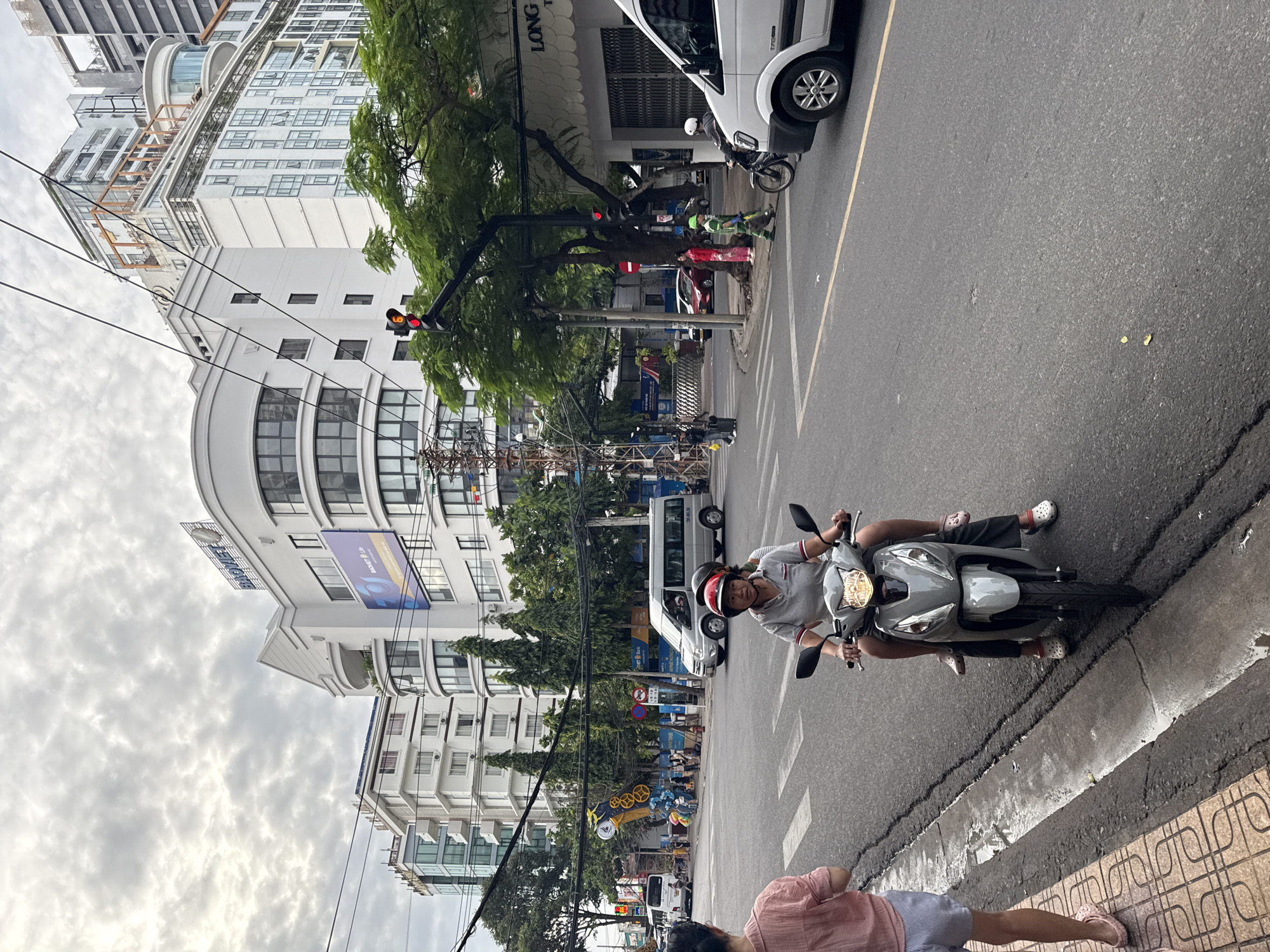 This image shows a street scene in Nha Trang, Vietnam. The foreground features a woman on a scooter wearing a red helmet, followed by another person, also on the scooter. To the left, a person wearing a pink shirt and striped shorts stands on the sidewalk. In the middle ground, a white van is parked on the right side of the road, and a silver van is in the middle of the intersection. Traffic lights are visible, with the red light illuminated. The background consists of modern buildings, including a curved white building with blue signage that reads "BAOVIET." Trees line the street, and utility wires are overhead. The sky is overcast.