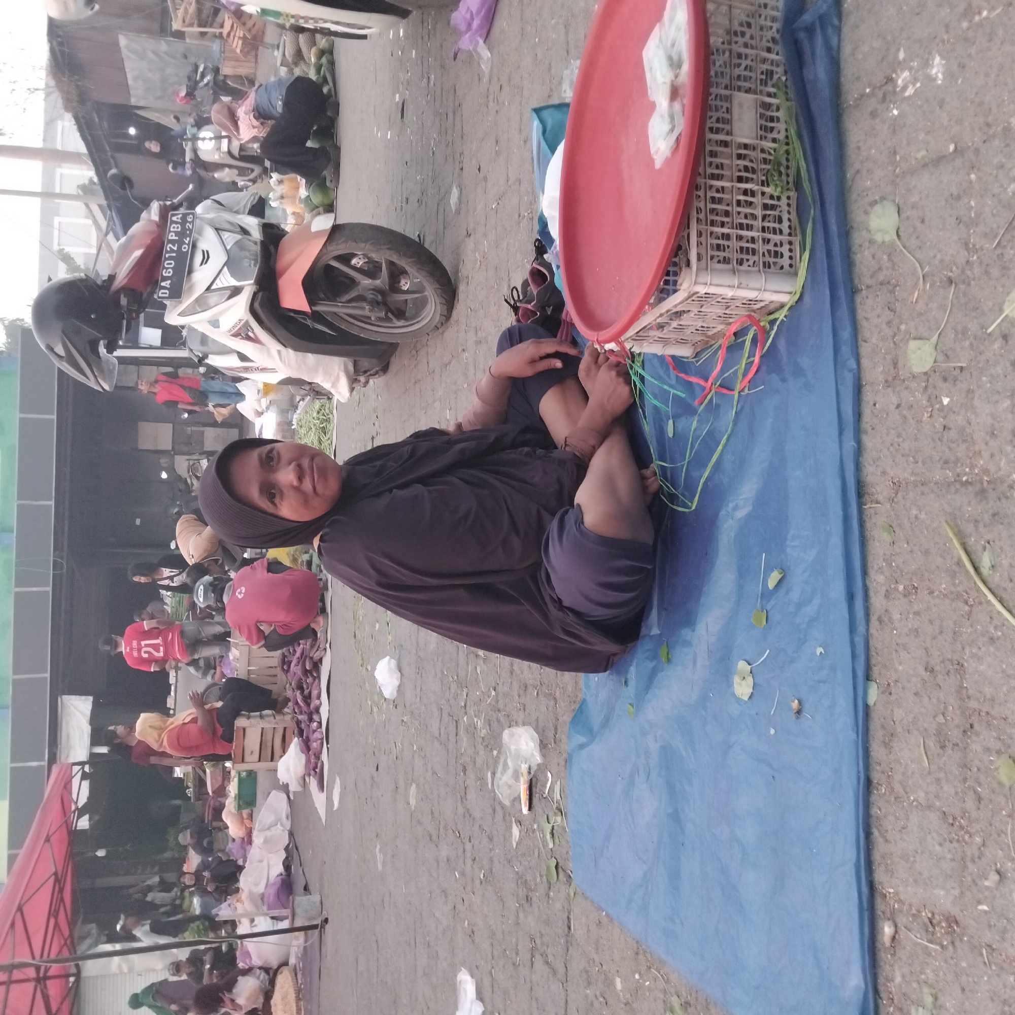 The image depicts an outdoor market scene, possibly in Banjarbaru, Indonesia. A woman wearing a dark hijab and black clothing is seated on a blue tarp on the ground. She is looking towards the camera. In front of her is a white plastic crate with a red circular lid on top, and what appear to be small packets of cash or goods on the lid. Several green and red strings are draped around the crate and tarp. Behind her, a white motorcycle with a black helmet on its handlebars is parked, and its license plate reads "DA 6012 PBA 04-26". Other market stalls and people are visible in the background, with some individuals appearing to be vendors or shoppers. Produce, such as eggplants, is also visible in the market. The ground is paved with asphalt or concrete, and there is some debris and fallen leaves scattered around. The overall impression is of a busy, everyday marketplace.