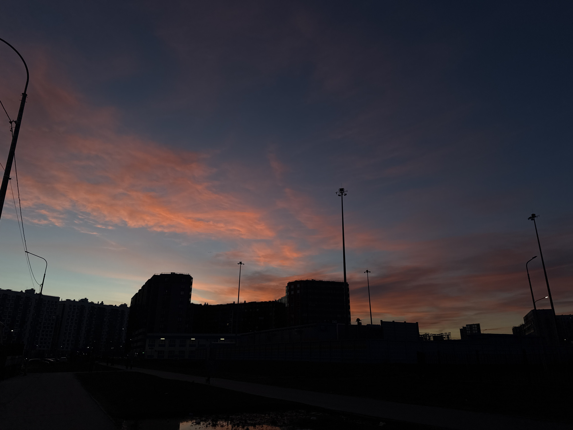 This image depicts an urban landscape at twilight, likely dusk, dominated by a vividly colored sky and dark silhouettes of city structures. The upper portion of the sky is a deep blue-grey, gradually transitioning into striking bands of orange, pink, and soft purple clouds that stretch across the horizon, illuminated by the low-angle sunlight.
In the foreground and midground, numerous multi-story buildings, appearing as dark, angular masses, are silhouetted against the vibrant sky. Their architectural details are largely obscured by shadow, though some faint window patterns can be discerned on the less dark sections. Several tall streetlights or lamp posts, some with multiple fixtures, rise prominently, also rendered in silhouette. A distinct lamp post with visible wires is positioned on the far left side of the frame.
A paved path or walkway extends across the lower part of the image, flanked by dark ground and indistinct vegetation. A small, reflective body of water or a large puddle is visible in the bottom foreground, subtly mirroring the ambient light. The scene appears quiet and still, with no discernible human activity or visible text. The architecture suggests a residential or urban development common in cities, fitting the location context of Russia. The overall mood is serene and dramatic, capturing the beauty of the fading light.