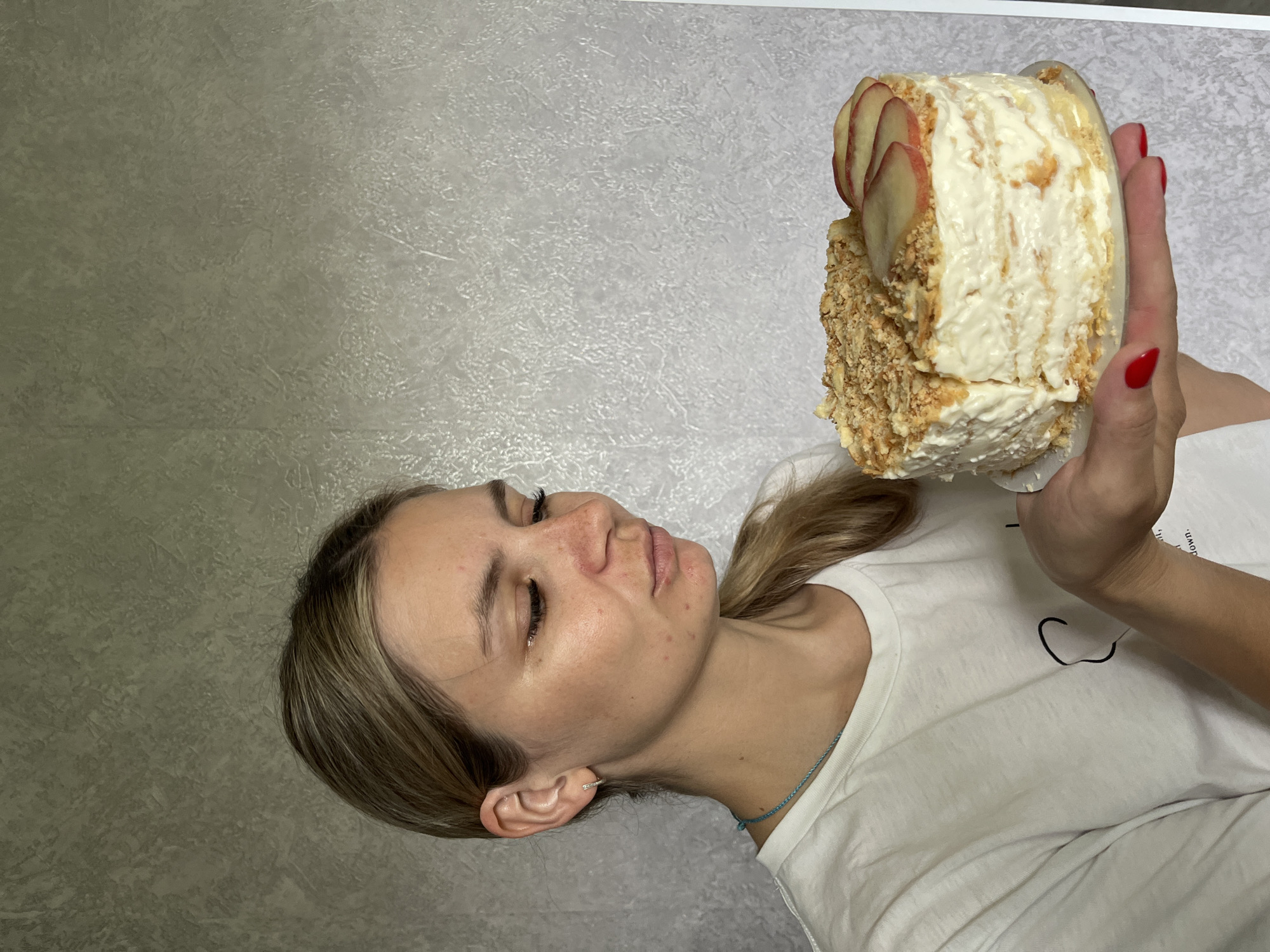 A young woman with blonde hair pulled back in a ponytail is holding a round cake with white frosting and sliced red apples on top. She is wearing a white t-shirt with a minimalist graphic design. The background is a textured grey wall. The image appears to be taken indoors. There is no indication of the specific location within Chaikovsky, Russia.