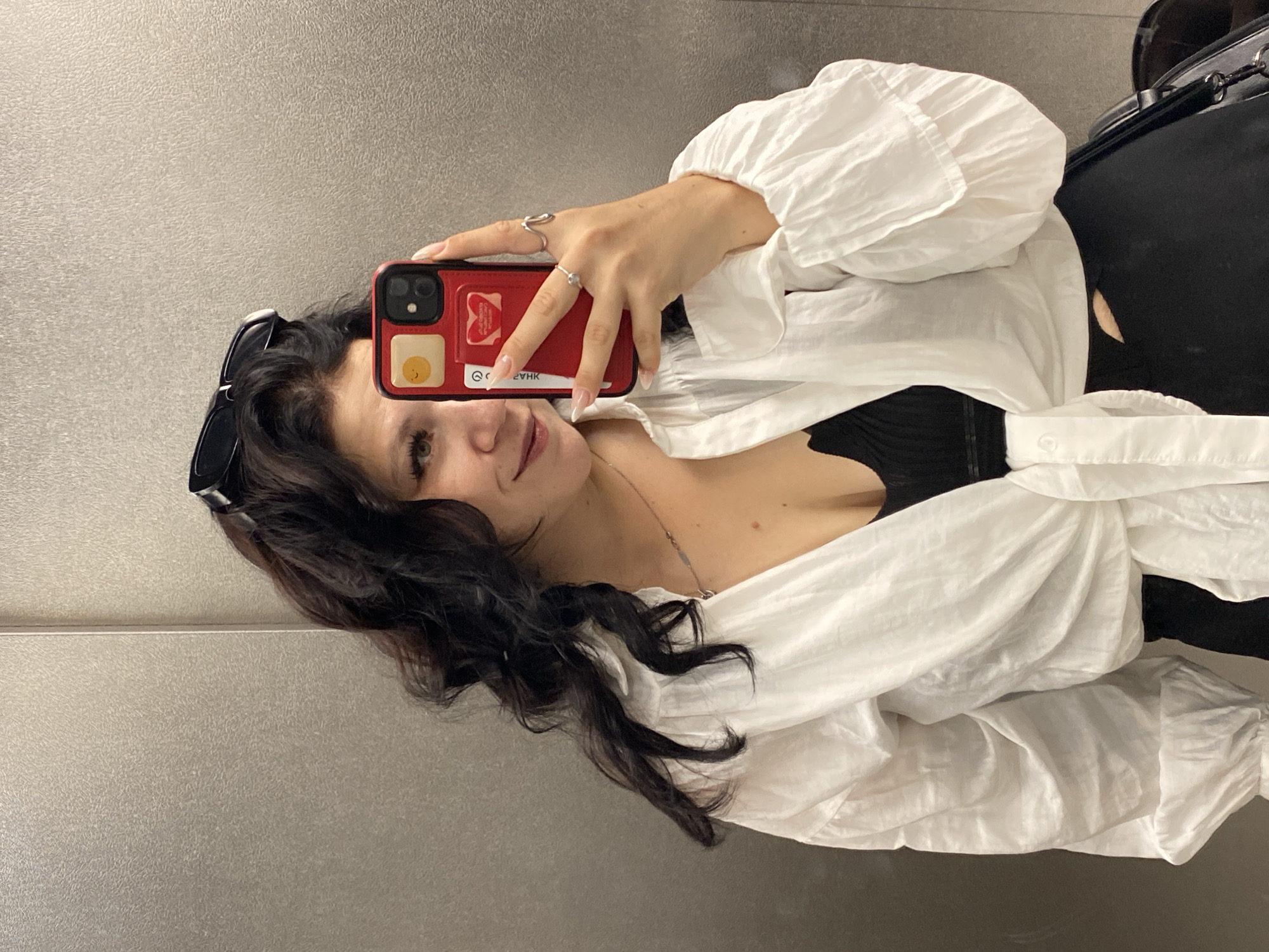A young woman with long, dark, curly hair is taking a selfie in what appears to be an elevator. She is wearing a white, tie-front blouse over a black top, and black pants. A pair of black sunglasses are perched on her head. She is holding a red phone in her right hand, and is wearing a couple of rings on her fingers. The elevator walls are a brushed metal texture.