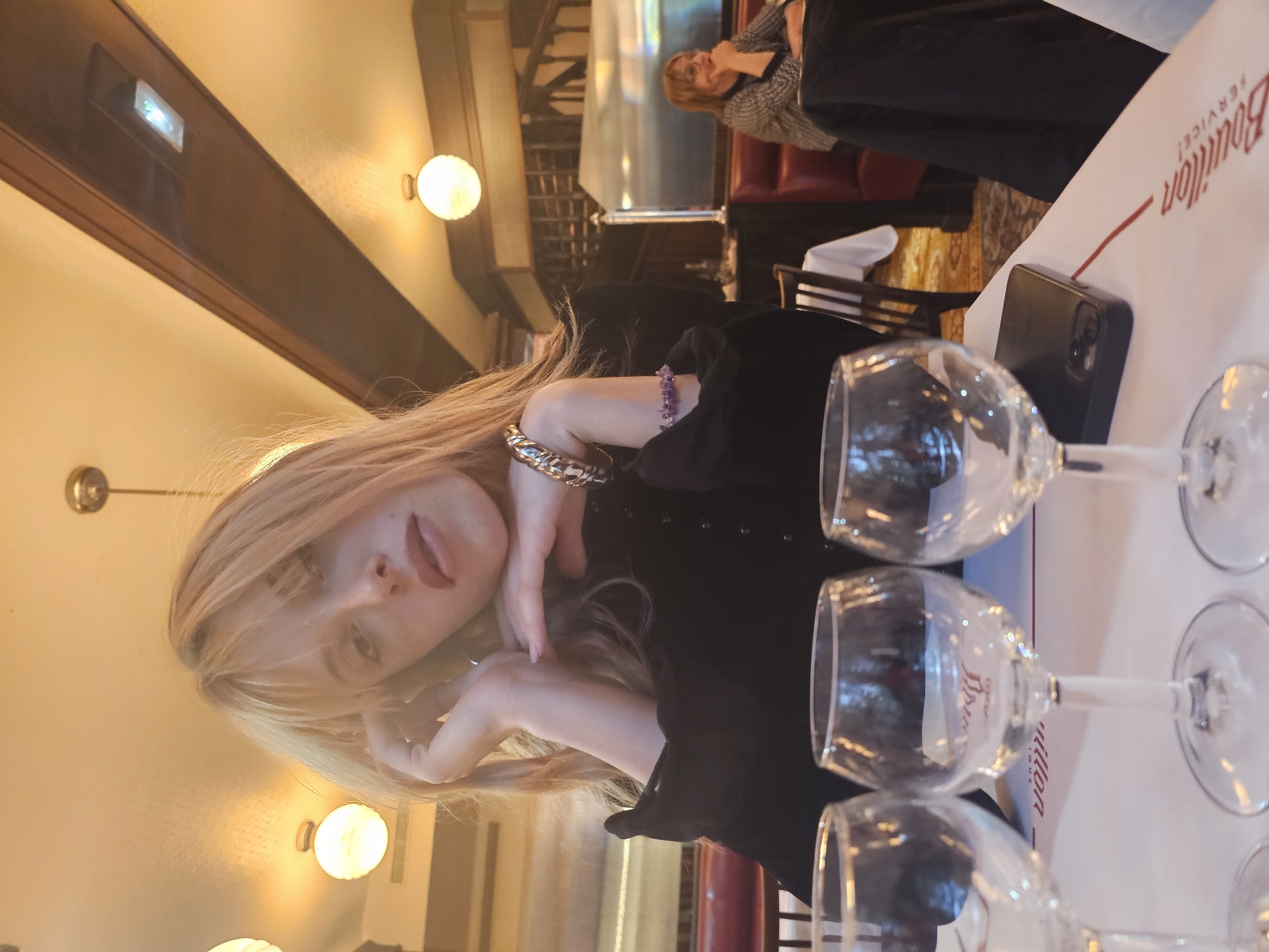 A young woman with blond hair is at a table in a restaurant. She is wearing a black top and has bracelets on her wrists. The restaurant appears to be the "Bouillon Service" restaurant, located in Paris, France, as evidenced by the document with that name visible in the shot. The table is set with wine glasses and a phone. Another woman is seated in the background. The lighting is warm and inviting, typical of a Parisian restaurant.