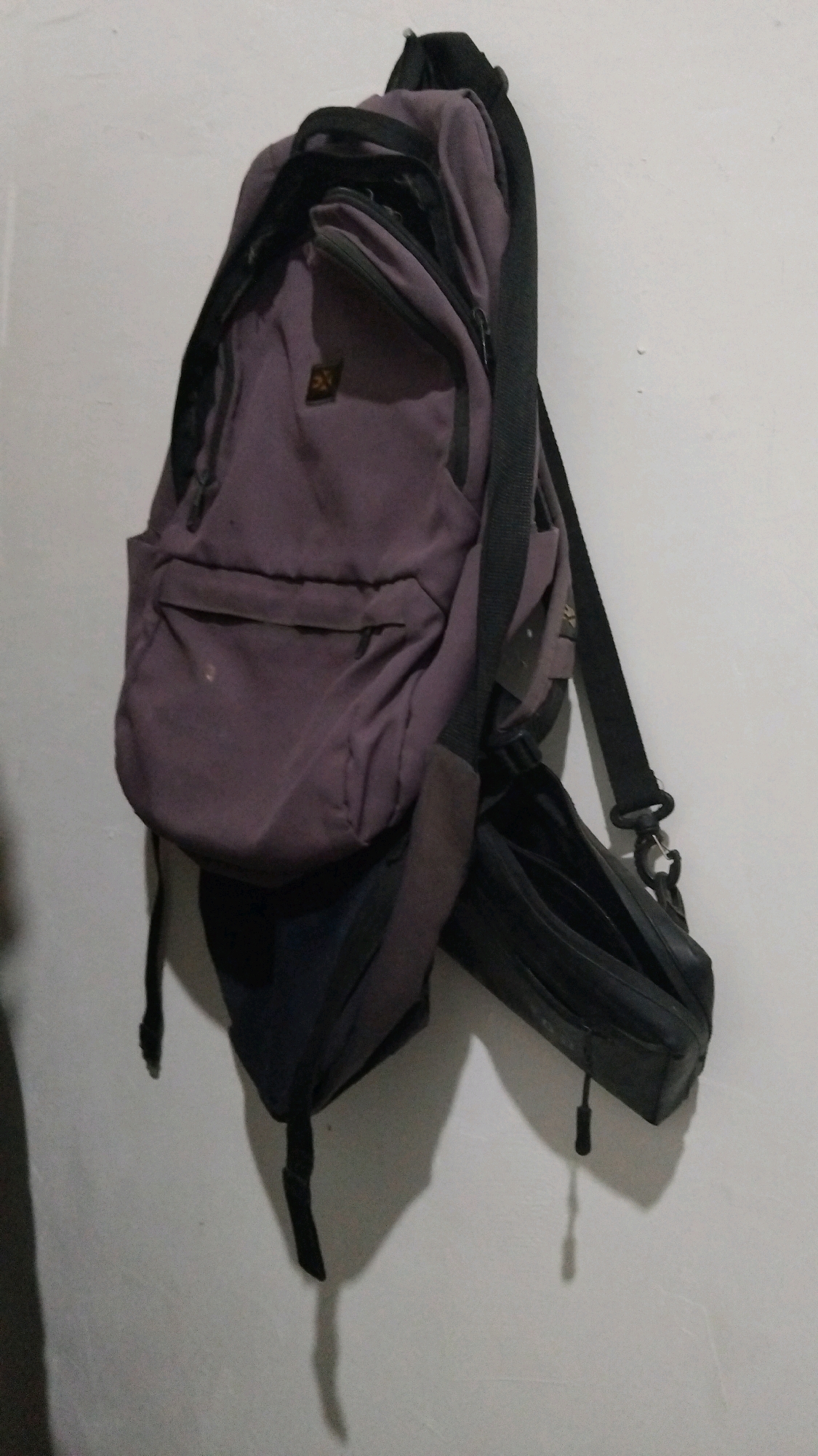 The image shows two bags hanging on a white wall. The bag on the left is a purple backpack with black straps. It has a front pocket with a zipper and a smaller pocket above it. The bag on the right is a black pouch or toiletry bag. Both bags are hanging from hooks or straps attached to the wall. The lighting is dim, casting shadows on the wall behind the bags. There are no people visible in the image. The location is described as Kebon IX, Indonesia, but this is not visually evident from the image itself.