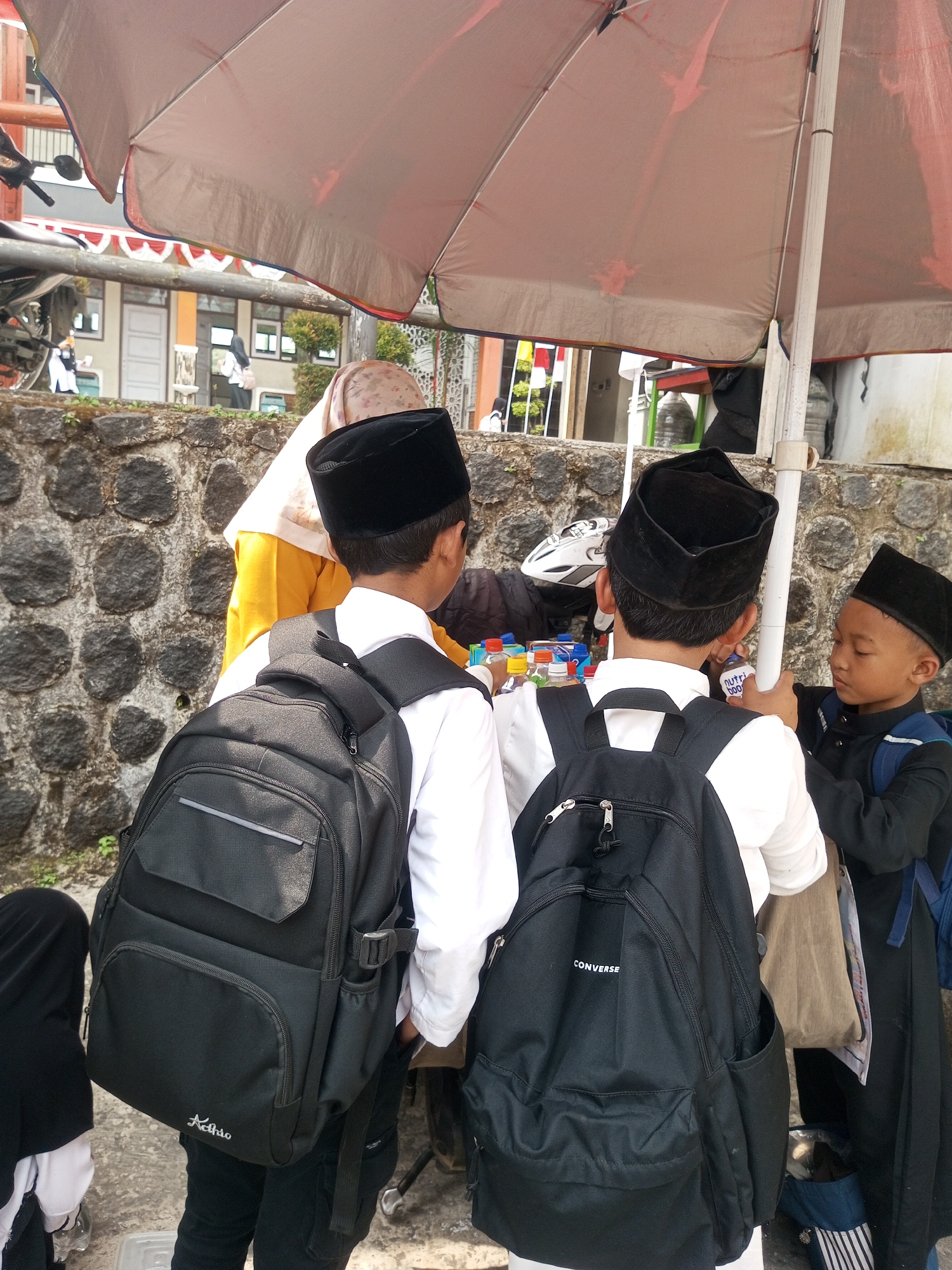 The image shows a street scene in Krasak, Indonesia, under a large, open umbrella that casts shadows. Three young boys, dressed in white shirts and black peci caps, are the central focus. Two of the boys are wearing large black backpacks. The boy on the left has a black backpack with a silver logo that reads "Achic" and appears to be looking down. The boy in the middle, also wearing a black backpack with "Converse" printed on it, is facing away from the camera and appears to be interacting with the boy on the right. The boy on the right, wearing a black outfit and peci, is looking at a bottle in his hand. In the background, a woman in a yellow shirt and headscarf is partially visible, along with a stone wall and a building with white and red trim. The ground is paved with concrete.