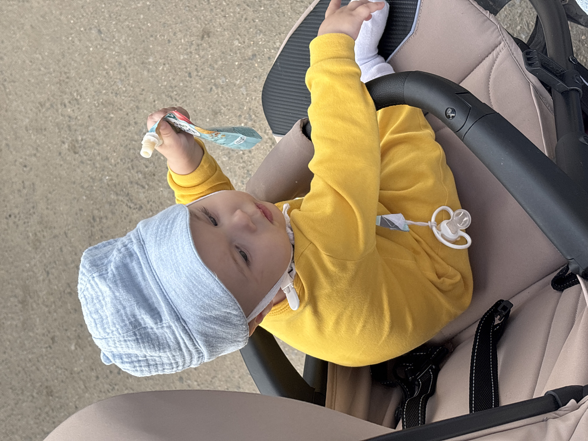 A toddler wearing a light blue hat and a yellow long-sleeved shirt is sitting in a stroller. The toddler is holding a pouch of baby food in their right hand. A pacifier is attached to the front of the toddler's shirt by a clip. The stroller is light brown with black straps and a black handle. The background is a gray, paved surface. The scene appears to be outdoors, possibly in a park or on a sidewalk. The location of Troshkovo, Russia is not discernible from the image.
