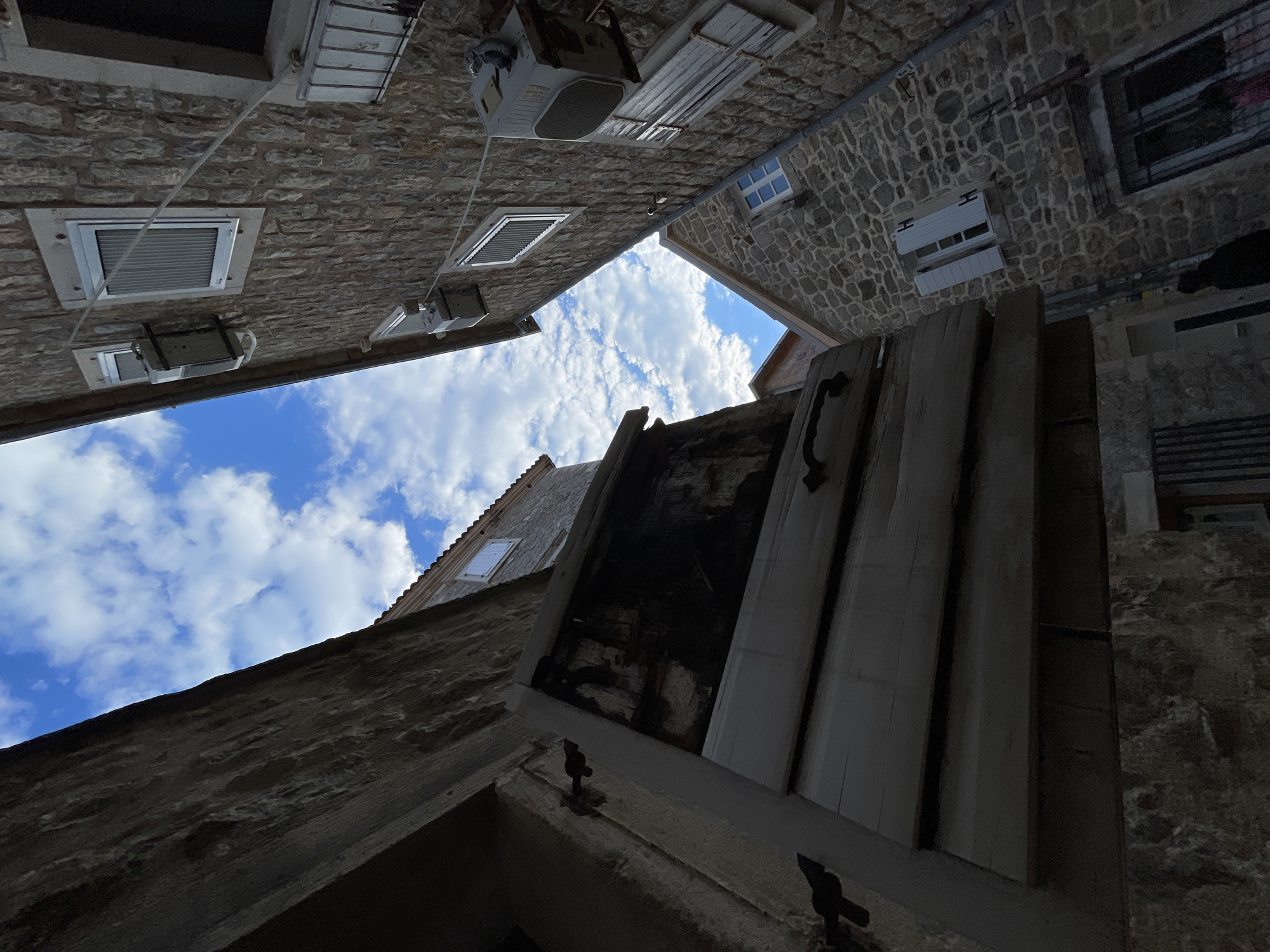 The image is a low-angle shot of a narrow alleyway between buildings in an unknown city. The buildings are constructed of stone, with several windows and air conditioning units visible on the walls. The sky is visible at the top of the frame, with white clouds against a blue background. In the foreground, a partially open wooden door with a black handle is visible. The perspective makes the buildings appear to tower over the viewer. The alleyway is shaded, creating a contrast with the bright sky.