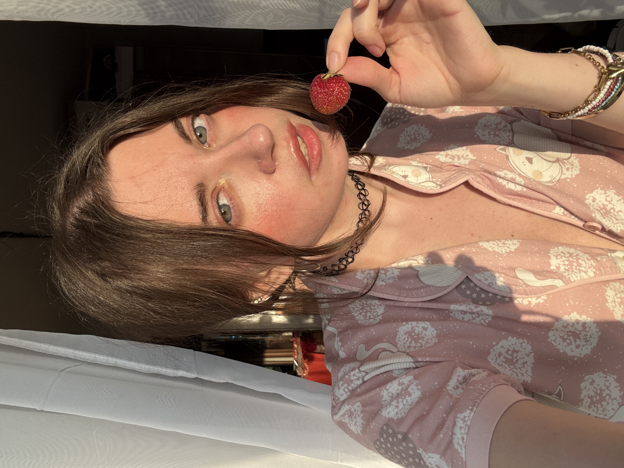 In this medium shot, a young woman is positioned in a vertical frame, her head tilted upwards and slightly to the left. She is holding a single strawberry between her thumb and index finger, bringing it close to her face. Her eyes, a striking blue-green, are looking upwards and slightly to the right, with a hint of golden shimmer on her eyelids. Her lips are slightly parted, revealing a hint of teeth, and her cheeks are flushed with a rosy hue, suggesting a playful or perhaps embarrassed expression. She wears a light pink pajama set adorned with a repeating pattern of white cartoon-like animals and floral motifs. A dark, thin choker necklace encircles her neck. On her left wrist, she wears a layered bracelet with beads of various colors and a gold chain. Her brown hair is loose and frames her face, with some strands falling across her forehead. The lighting suggests it is late afternoon or early morning, with warm sunlight casting shadows and highlighting her skin. She appears to be indoors, possibly near a window, as indicated by the white sheer curtains in the foreground and the soft light. The background is blurred, showing hints of a bookshelf and dark surfaces.