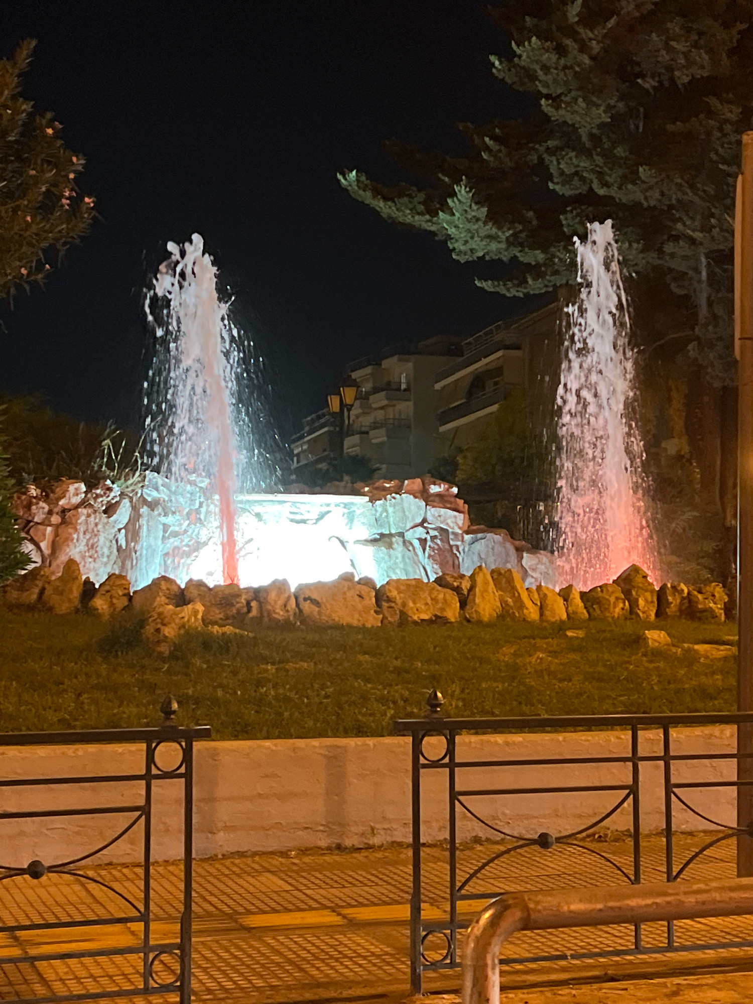 This is a night-time, eye-level, full shot of an illuminated fountain in the city of Kallithea, Greece. The fountain has two prominent water jets spraying upwards, one on the left and one on the right. The water is illuminated with pink and white light, creating a dynamic visual effect. The fountain itself is surrounded by a rock formation that is also lit from within, casting a bright glow. In front of the fountain is a patch of grass. Behind the fountain, buildings can be seen, suggesting an urban setting. A street lamp is visible on the right side of the image. In the foreground, a metal fence with a decorative pattern separates the viewer from the fountain area. The ground in front of the fence is paved and shows a pattern of light and shadow. The overall atmosphere is one of a well-maintained public space, likely a park or a plaza, illuminated for evening enjoyment.