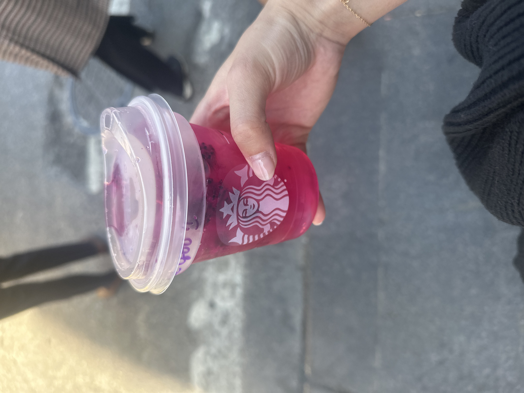 A person holds a Starbucks Dragonfruit Refresher in New York City. The drink is in a clear plastic cup with a translucent lid. The Starbucks logo is visible on the side of the cup. The person is wearing a black sweater and a gold bracelet. The background is blurry and shows a gray sidewalk.