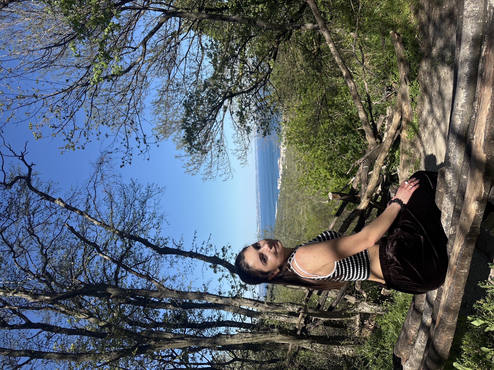 A medium shot shows a young woman sitting on a wooden structure within a forested area. The setting is likely in or near the city of Varna, Bulgaria, as the sea and a coastline are visible in the background through the trees. The woman is wearing a striped tank top and dark velvet pants. She is facing the camera with a slight smile. The forest consists of deciduous trees with thin branches, some of which are bare while others have small green leaves. The sky is a clear blue. The wooden structure appears to be a makeshift fence or seating area made from logs. Green vegetation grows around the structure.