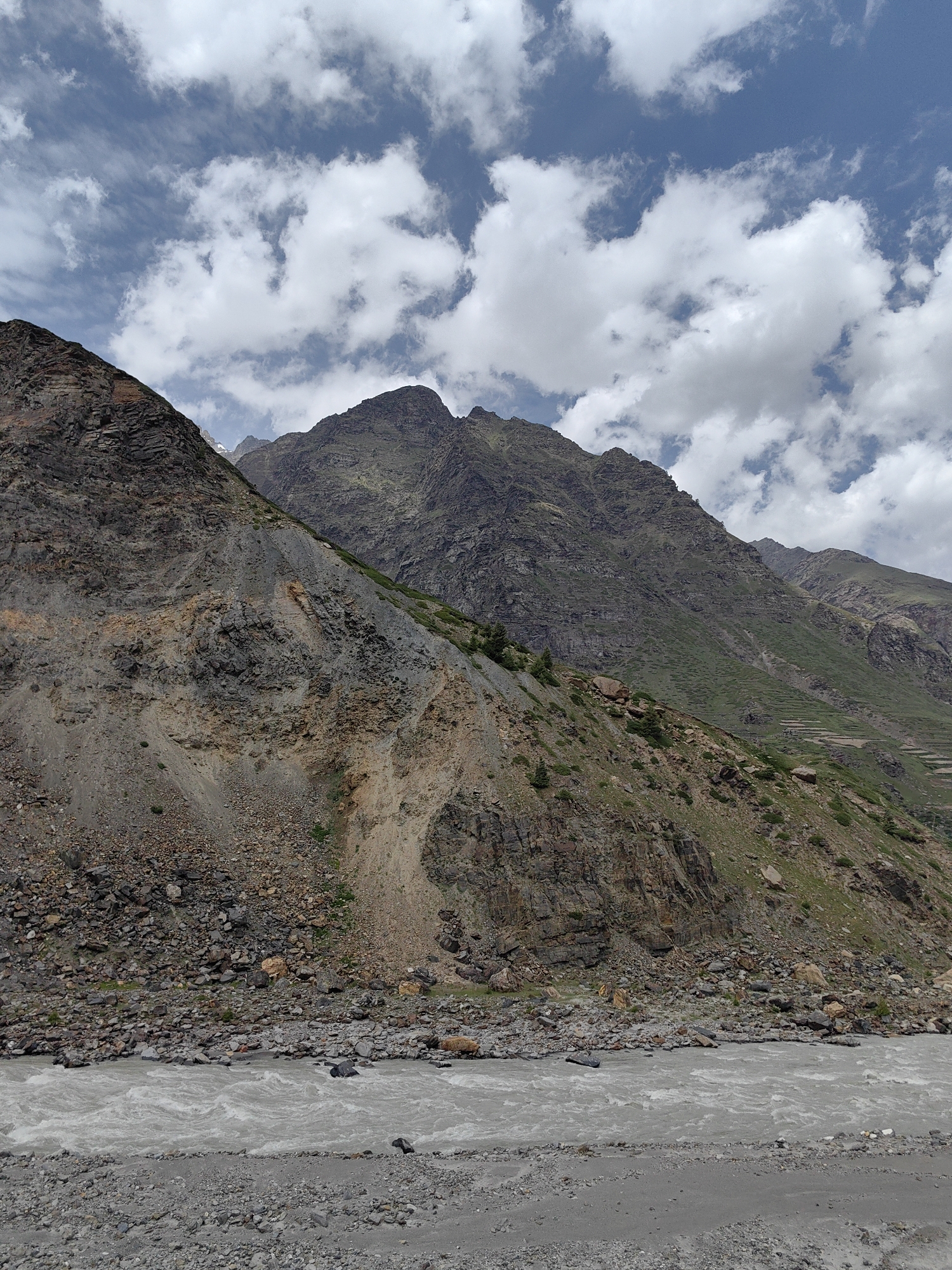 The image captures a vast, mountainous landscape in Sissu, India, dominated by rugged, rocky slopes and a turbulent river. The foreground features a grey, gravelly riverbed with the shallow, fast-flowing river snaking through it. The water appears murky, indicating it carries sediment.
Behind the river, the mountains rise steeply. The left side of the image showcases a stark, eroded mountainside with exposed layers of rock and scree, displaying shades of brown, grey, and ochre. A few scattered, hardy green plants cling to the lower slopes.
The right side of the mountain range is covered in a patchwork of green vegetation, interspersed with rocky outcrops. In the mid-ground, terraces carved into the mountainside suggest human agricultural activity, with a few small structures visible.
The sky above is a bright blue, scattered with dramatic, white cumulus clouds that cast shadows across the mountain peaks. The overall scene is one of wild, untamed natural beauty, typical of high-altitude mountainous regions. There are no people or animals visible in the frame.