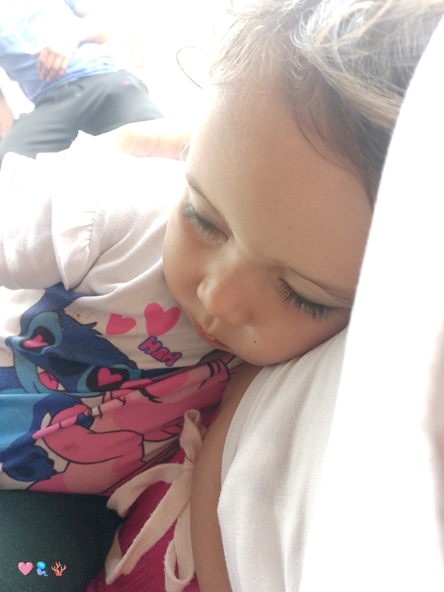 A close-up shows a sleeping child, resting against a person's shoulder covered by a white garment. The child wears a white t-shirt with a cartoon print featuring pink hearts and a blue character. Part of an adult's body, wearing a blue shirt and gray pants, is visible in the blurred background.
The scene's location in El Cercado, Venezuela, cannot be determined from the image.