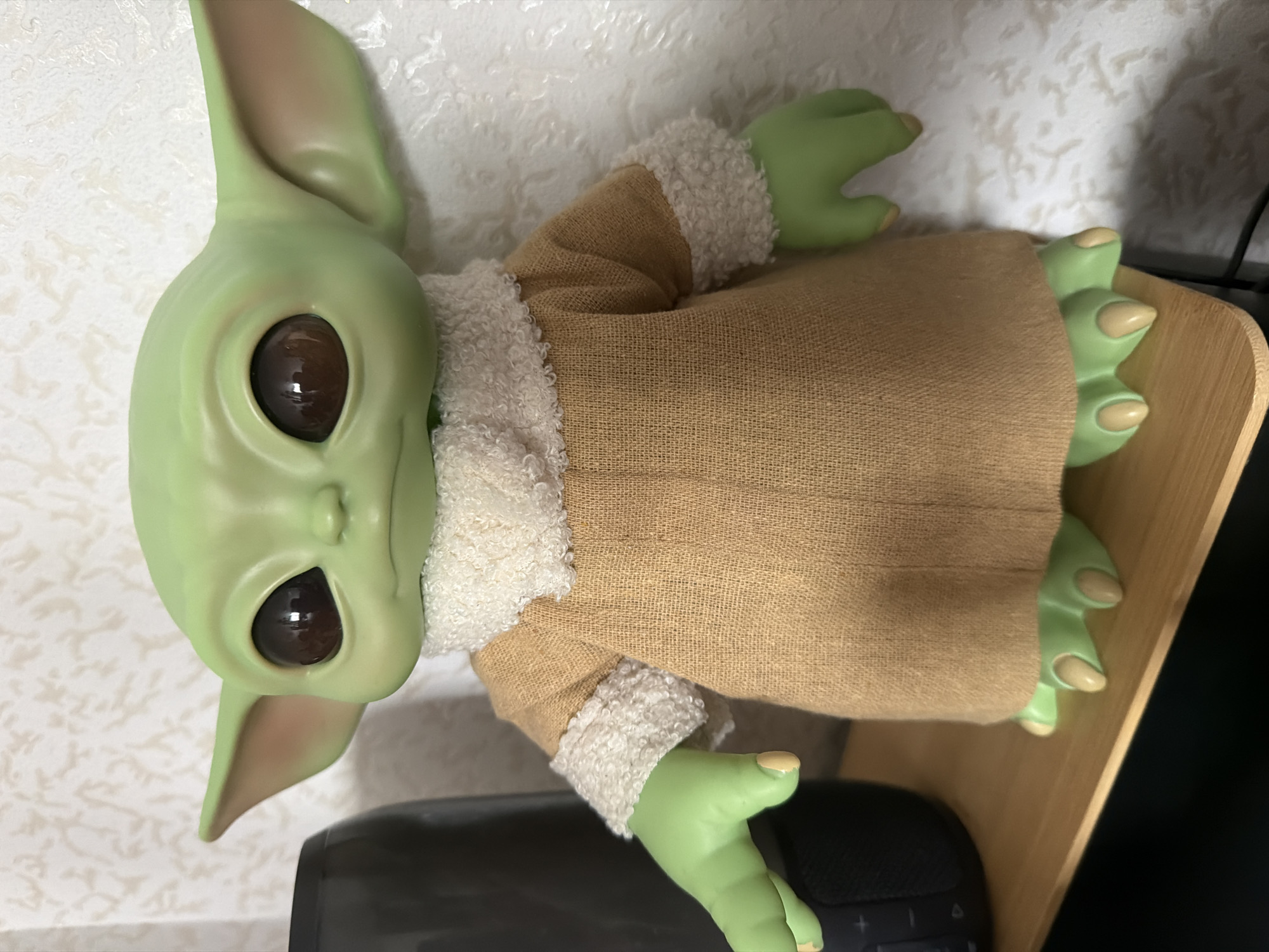 A doll resembling the character "Baby Yoda" is positioned on a wooden shelf. The doll has large, dark eyes, green skin, and is wearing a tan robe with a white, furry trim around the neck and sleeves. Its feet are visible at the bottom, also green with tan tips. To the left of the doll, a dark, cylindrical object, possibly a speaker, is partially visible. The background consists of a white, textured wall. The setting is indoors, with no specific details to indicate a location in Yablonovskiy, Russia.