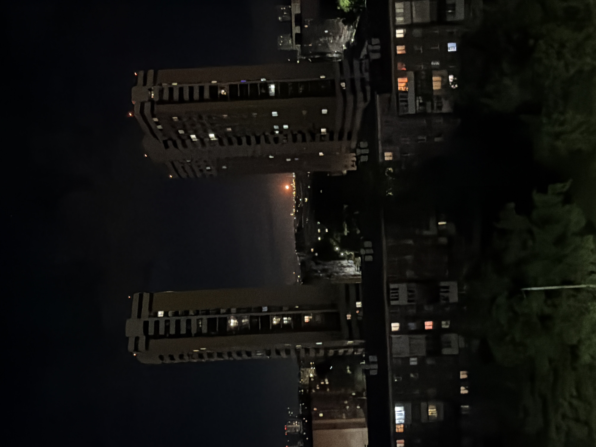 The image shows a night view of a city with multiple buildings. Two tall buildings with multiple lit windows are prominent on the left side of the image. Other buildings with illuminated windows are also visible. Some dark trees are seen on the right side of the image. A faint orange glow is on the horizon. The location is in a city, but specific location details are not clear from the image.