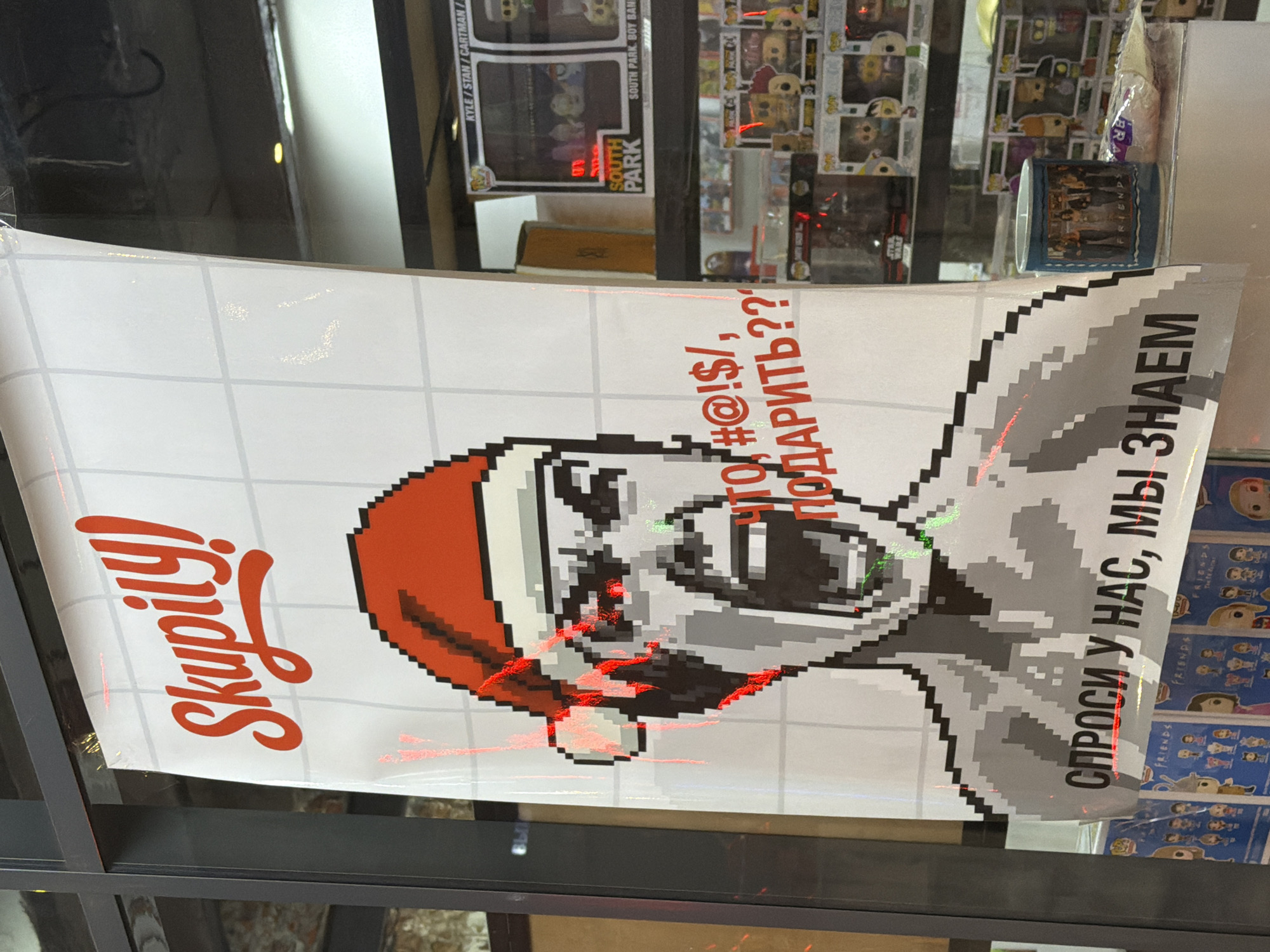 This is a photograph of a poster displayed in a shop window in Saint Petersburg, Russia. The poster features a pixelated image of a person wearing a Santa hat with an angry, yelling expression and red, glowing eyes. The text on the poster is in Russian. The top of the poster reads "Skupliy!" which translates to "Stingy!". Below the face, it says "ЧТО, #@!$, ПОДАРИТЬ??" which translates to "What, #@!$, to give as a gift??". At the bottom of the poster, it reads "СПРОСИ У НАС, МЫ ЗНАЕМ" which translates to "Ask us, we know". The poster appears to be for a gift shop, likely related to the Christmas season. Behind the poster, the interior of the shop is visible, containing shelves filled with merchandise, including "Funko Pop" collectible figures. The shop window is framed by dark metal.