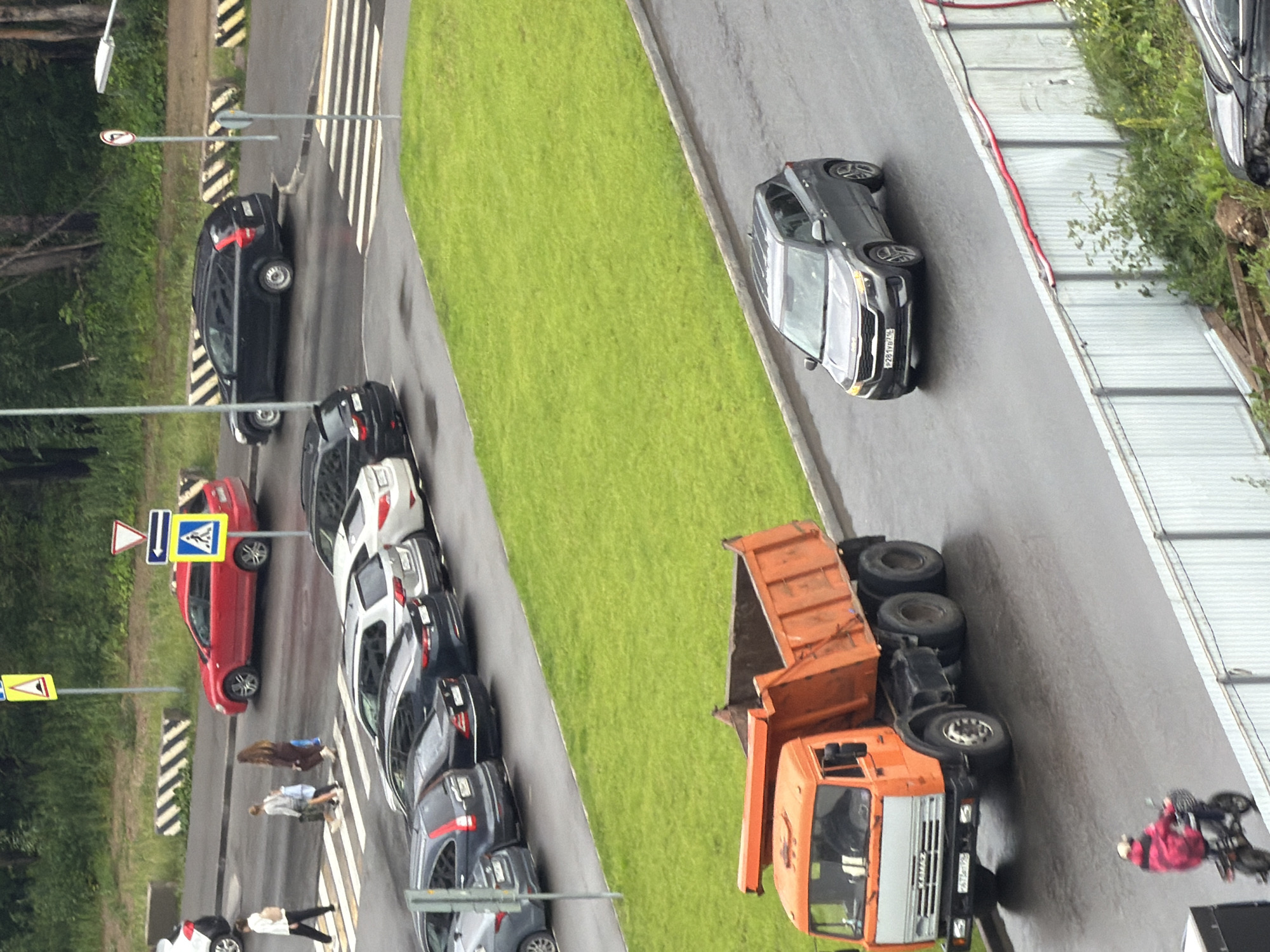 In an urban environment, possibly a city, a grey SUV is driving on a wet asphalt road. The SUV has a license plate that reads "P281VB774". To the left of the SUV, a large orange dump truck is parked on the road, with its bed raised. The dump truck has a license plate with the characters "P673TX". In the background, there is a patch of green grass bordered by a concrete curb, and behind that, more trees and foliage. Further in the distance, several cars are parked and driving on what appears to be a street with crosswalks. A few pedestrians are visible on the crosswalk. The scene is observed from an elevated vantage point.