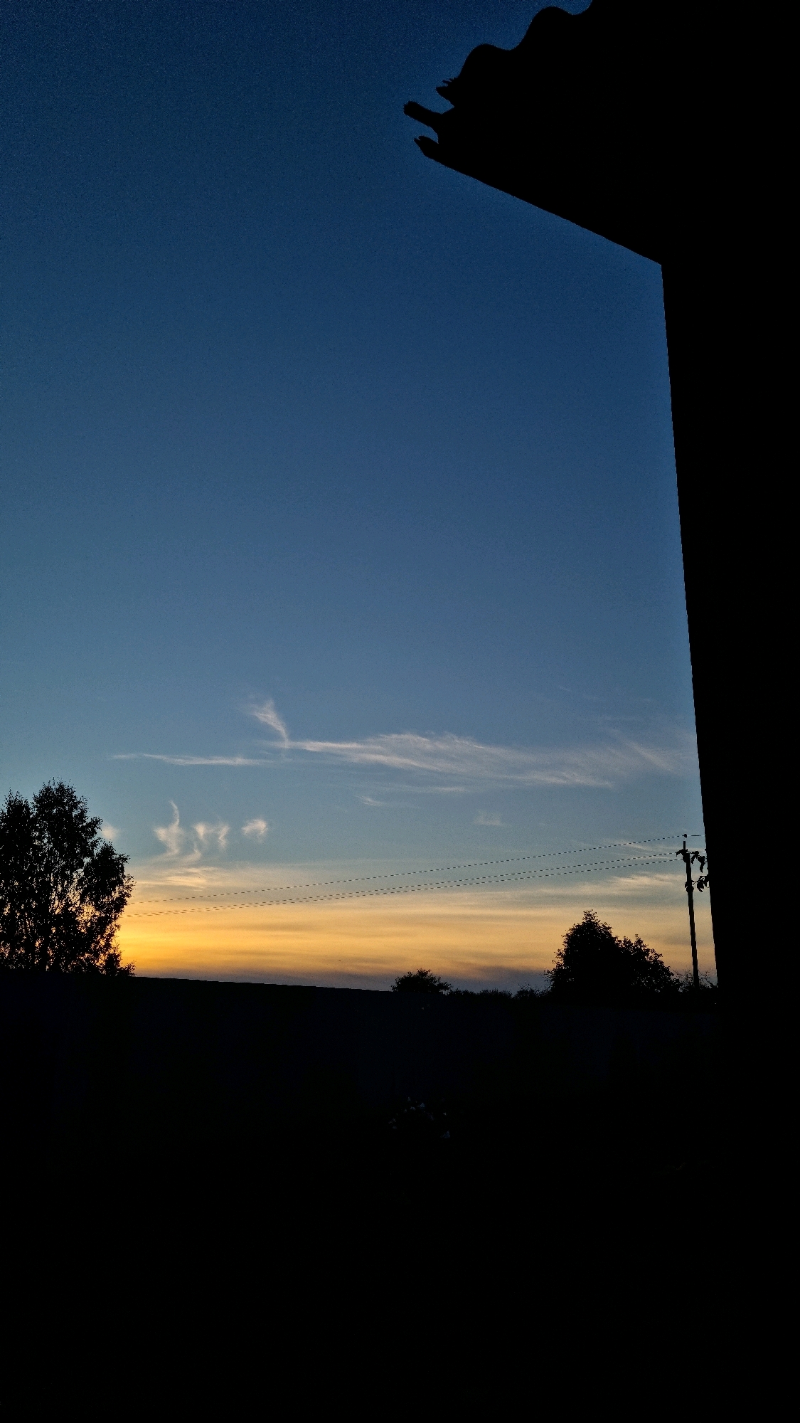The image shows a sunset scene with a dark silhouette of trees and a field against a gradient sky of deep blue, fading to orange and yellow near the horizon. A dark, angular structure, possibly a building or roofline, is visible in the upper right corner. Several thin, wispy clouds are scattered across the sky, with two power lines running horizontally through the mid-ground. A solitary utility pole with insulators is situated on the right side of the frame. The overall impression is one of a rural or semi-rural setting at dusk, with a distinct lack of human presence or activity. The location is Vysokoe, Russia.