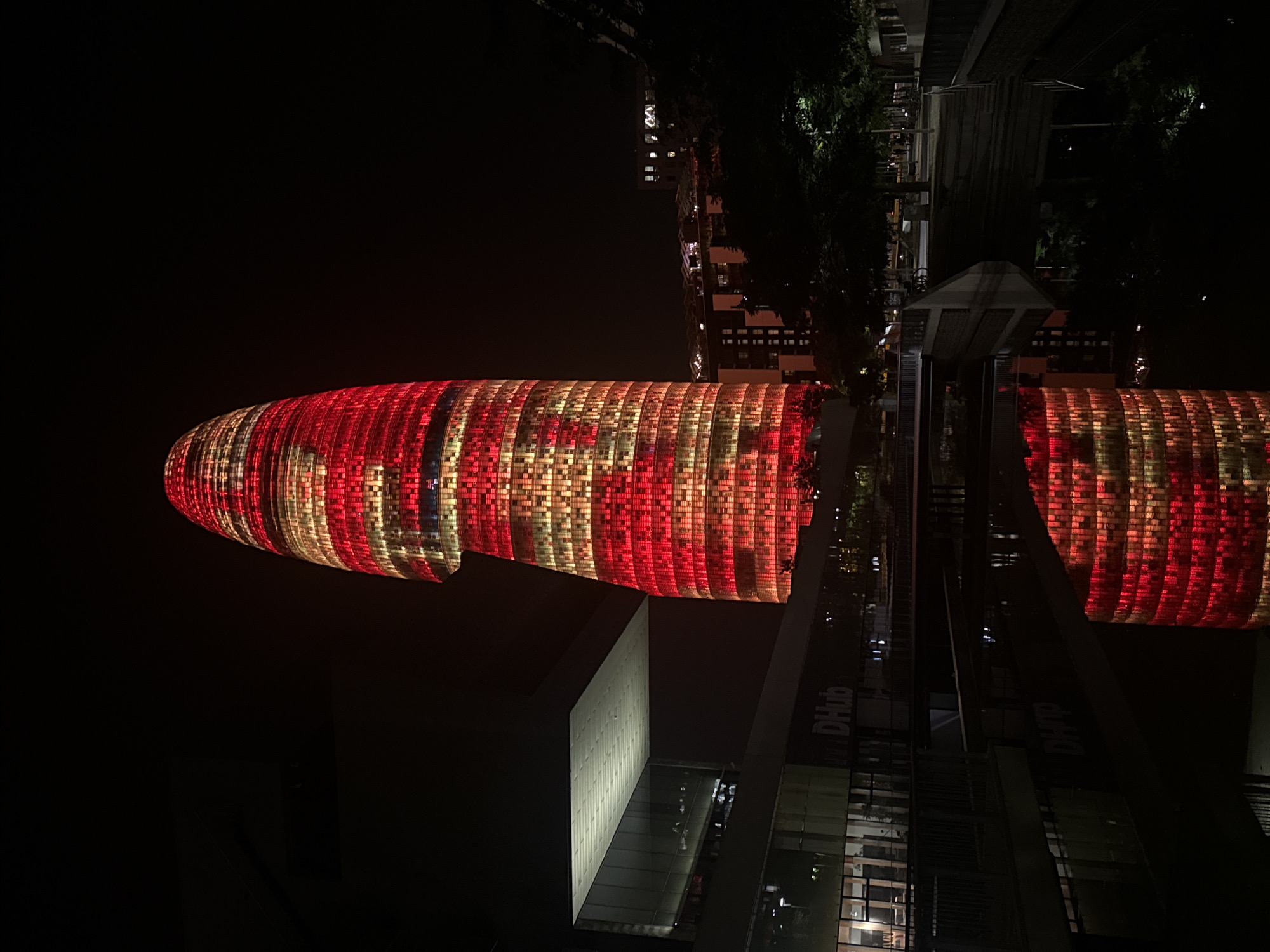 This is a night-time, low-angle, full shot of the iconic Agbar Tower in Barcelona, Spain, illuminated with red and yellow lights in a horizontal striped pattern, with some sections appearing to be a solid red. The tower's distinctive bullet shape is prominent, its top reaching into the dark sky. In the foreground, a body of water reflects the illuminated tower. To the left of the tower, a modern building with a large, angled, illuminated white roof and glass facade is visible. The text "DHUB" is visible on this building. The environment suggests an urban setting with trees and other buildings visible in the background, also partially illuminated. The scene is well-lit on the tower and the nearby building, creating a strong contrast with the dark night sky and surrounding areas. There are no people clearly visible in the image.