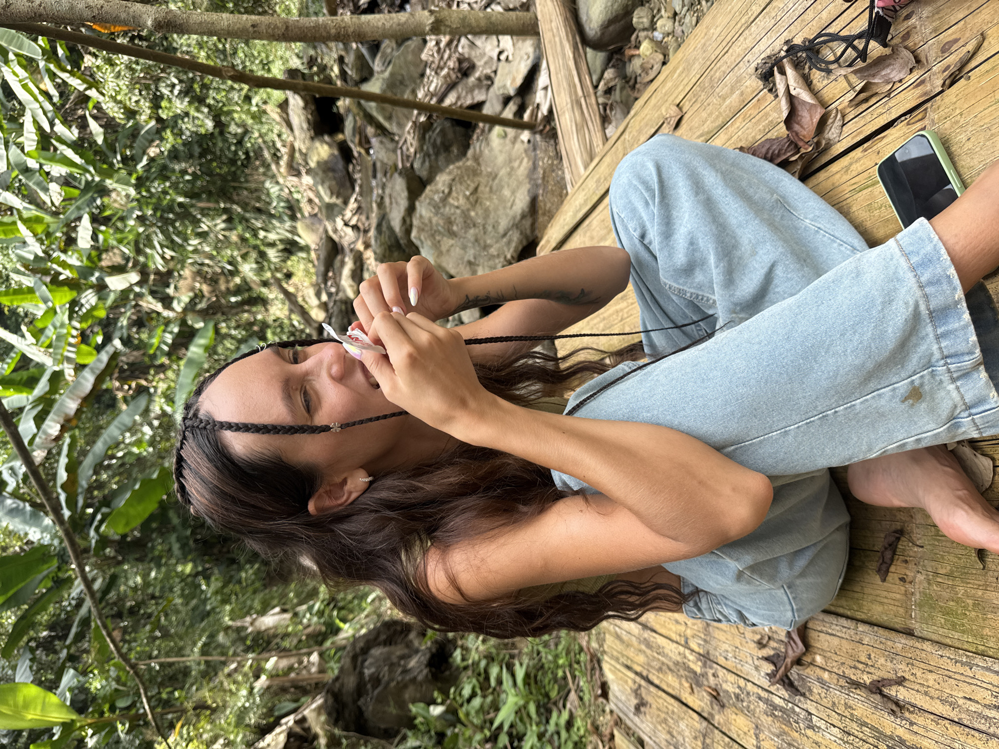 A young woman is sitting cross-legged on a bamboo platform in Suối Giàng, Vietnam. She is wearing light blue jeans and has long brown hair with braids. She is smiling and holding a small white object in her hands, possibly applying makeup. A smartphone with a green case sits nearby on the platform. The background includes lush greenery, rocks, and a small stream.