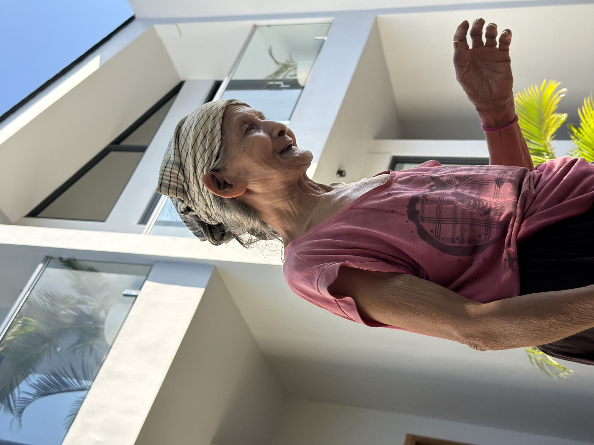 An elderly woman with graying hair tied up in a patterned headscarf stands outdoors, looking down and to her right with a slight smile. She is wearing a pink t-shirt with a graphic design and a dark skirt. Her left arm is bent at the elbow, and her right hand is extended forward, palm up, with fingers slightly curled. Behind her, a modern white building with glass balconies and a clear blue sky is visible. The scene appears to be in Kuta Selatan, Indonesia, suggested by the tropical foliage in the foreground and the architectural style.