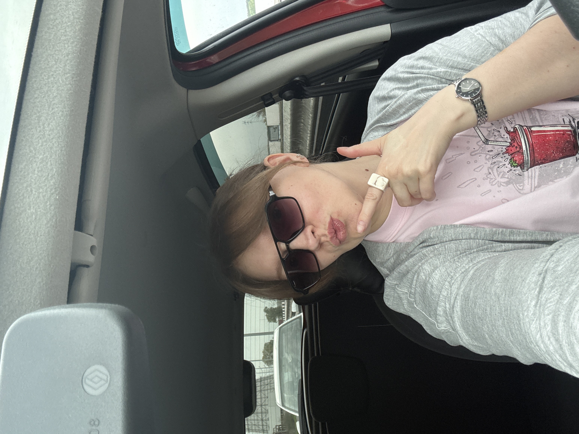 A woman sits in the driver's seat of a vehicle, wearing sunglasses, a light pink t-shirt with a strawberry smoothie graphic, and a grey cardigan. She has a metal watch on her left wrist and a large white ring on her right index finger. The woman is making a "duck face" expression. The vehicle is parked in a location that appears to be a parking lot or industrial area, possibly Izhevsk, Russia, based on the “Renault” logo visible on the rear view mirror. A white van can be seen through the vehicle window. The interior of the vehicle is visible.