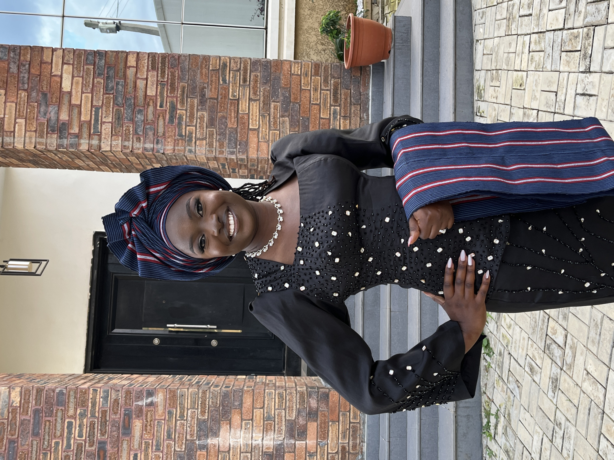 A dark-skinned woman with braids wears a black, embellished dress and a matching headwrap in front of a brick building. She is smiling at the camera, with her left hand on her hip and her right arm bent. She is holding a striped blue, white, and red cloth. The building has a dark door and a set of stairs leading up to it. A plant in an orange pot is to the right of the stairs. The ground is paved with light-colored tiles. The sky is cloudy.