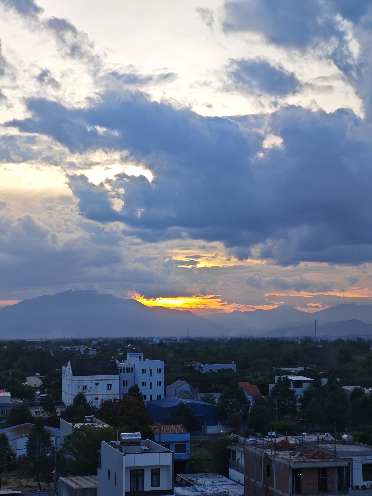 This is a vertical, medium shot of a dusky sky over a city landscape in Da Nang, Vietnam. The sky is dominated by dramatic clouds, with patches of golden light breaking through, suggesting either sunrise or sunset. Below the sky, a mountain range is silhouetted against the horizon. The city below is a mix of buildings, including what appears to be a white, multi-story building with many windows, and other, smaller structures interspersed with dense greenery. The overall atmosphere is one of impending twilight.