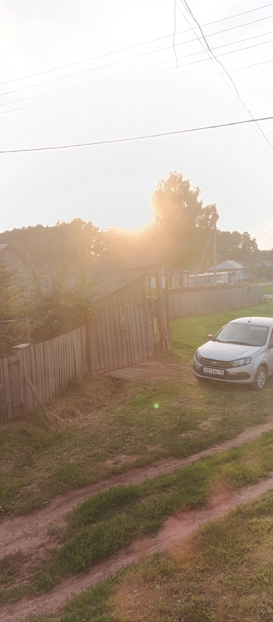 The image captures a sun-drenched scene in Podgornoye, Russia. In the foreground, a dirt road with tire tracks winds through overgrown grass. To the right, a silver sedan is parked on the grass. Behind the car, a wooden fence encloses a grassy area with a rustic wooden structure, possibly a shed or small building with a sloped roof. Further back, houses and trees are visible, with the bright sun casting a strong glare, partially obscuring the details of the sky and distant landscape. Power lines stretch across the upper portion of the frame.