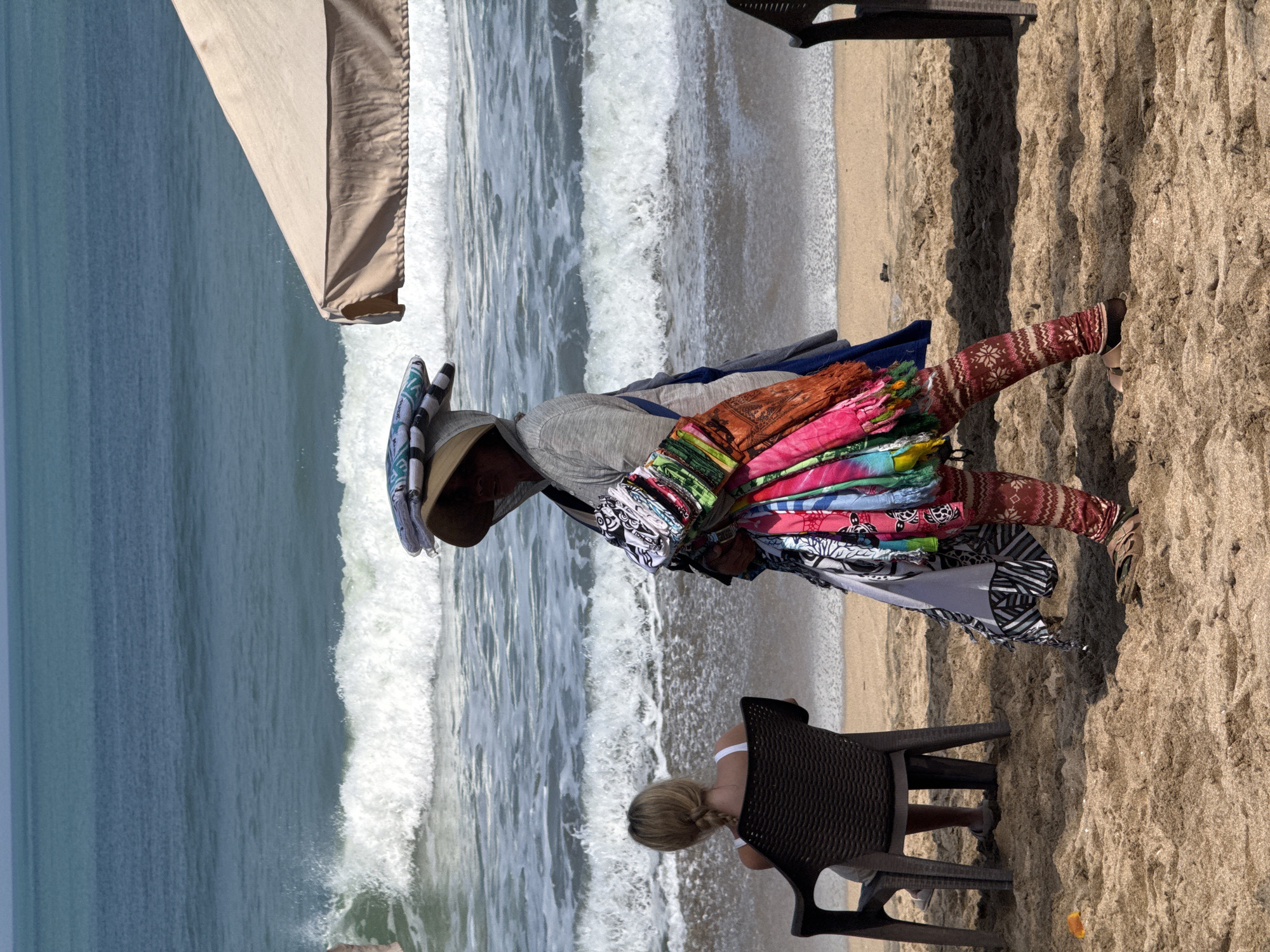 A person is walking on the beach in Kuta, Indonesia, carrying a stack of colorful fabrics on their head and a bundle of textiles in their arms. They are wearing patterned leggings and sandals. To their left, a woman with braided blonde hair sits in a dark plastic chair, facing the ocean. Waves crash on the sandy shore behind them. A light-colored beach umbrella is partially visible on the right side of the frame. The sky is clear and the water is a deep blue.