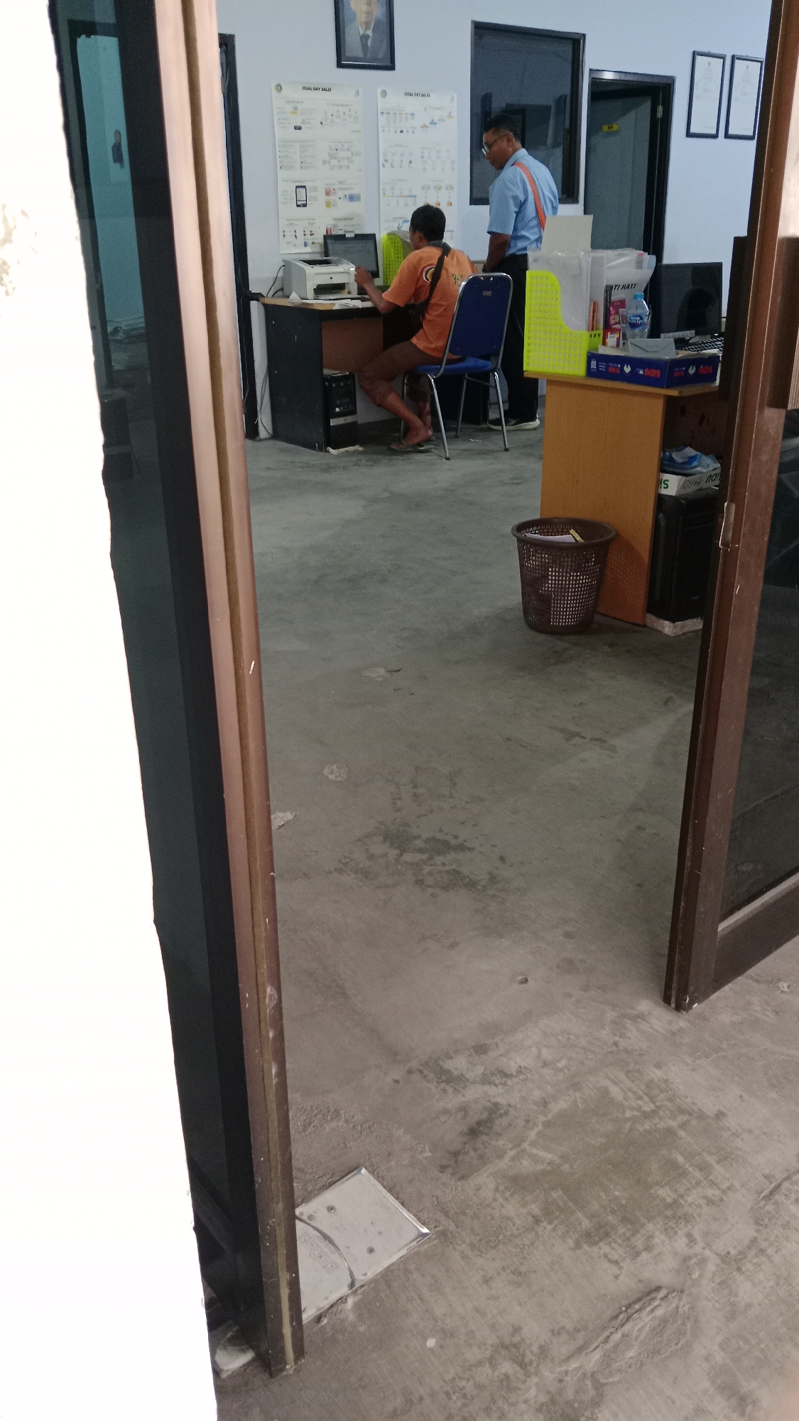 In Blitar, Indonesia, a man in an orange shirt sits at a desk with a printer and laptop, facing a bulletin board with diagrams. Another man in a blue shirt with an orange stripe stands nearby, observing. The scene appears to be an office or training room, with a trash can, filing baskets, and documents on a desk. The flooring is concrete, and there is a doorway leading to another area.